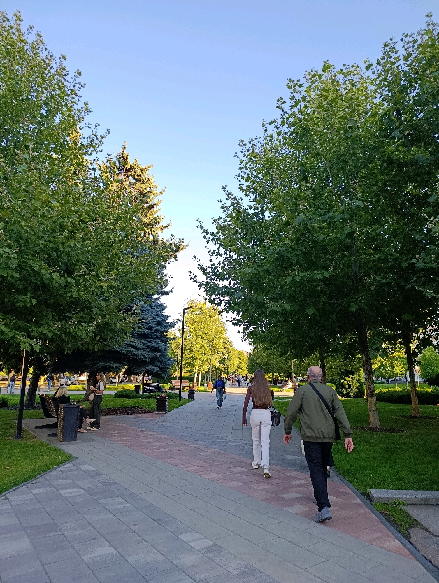The image depicts a sunny day in a park-like area in Cherkasy, Ukraine. A paved pathway, constructed with grey and pinkish-red bricks, runs through the scene. Lush green trees with dense foliage line both sides of the pathway, creating a shaded atmosphere. In the foreground, two individuals are walking away from the camera on the pathway. A man in a green jacket and dark pants walks ahead, while a woman in white pants and a brown top follows behind. Further down the path, other individuals are visible, some walking and some seated on benches. One bench, located to the left of the pathway, is occupied by two women. A trash bin is situated next to the bench. The sky above is a clear, light blue. The overall impression is that of a well-maintained public space, likely a boulevard or park, enjoyed by people during pleasant weather.