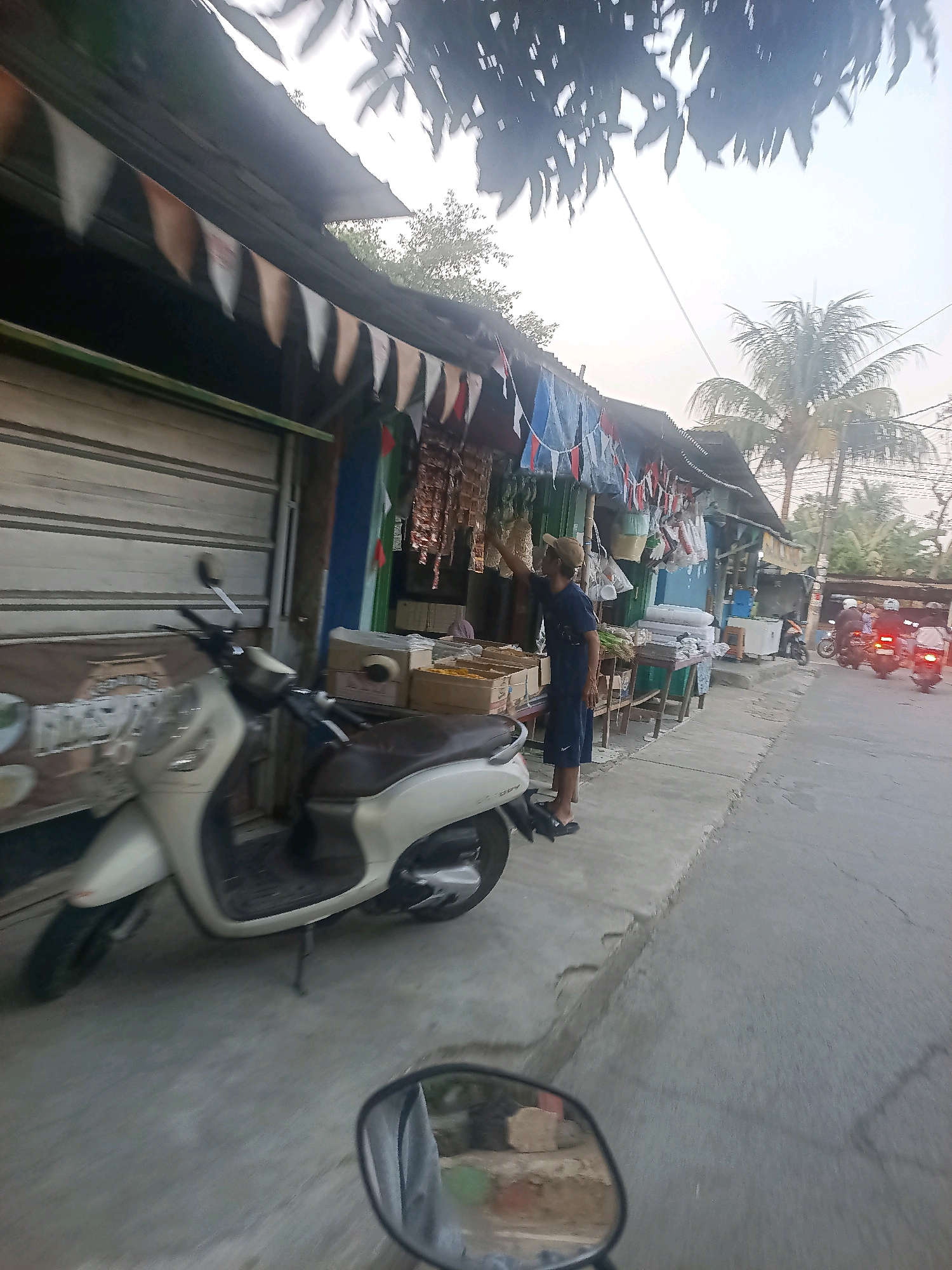 The image depicts a street scene in Telagaasih, Indonesia, with a focus on a small roadside stall. A light-colored scooter is parked on the left side of the frame, partially obscuring a closed storefront with a metal roll-up door. A man wearing a dark t-shirt and shorts stands at the stall, which is adorned with small flags or bunting. The stall displays various goods, including what appear to be packaged food items hanging from the awning, and boxes of merchandise on tables. In the background, several motorcycles are being ridden on the street, with their headlights illuminated. Palm trees are visible in the distance, and the sky is overcast. The scene suggests a typical day at a local market or shop in an Indonesian town.