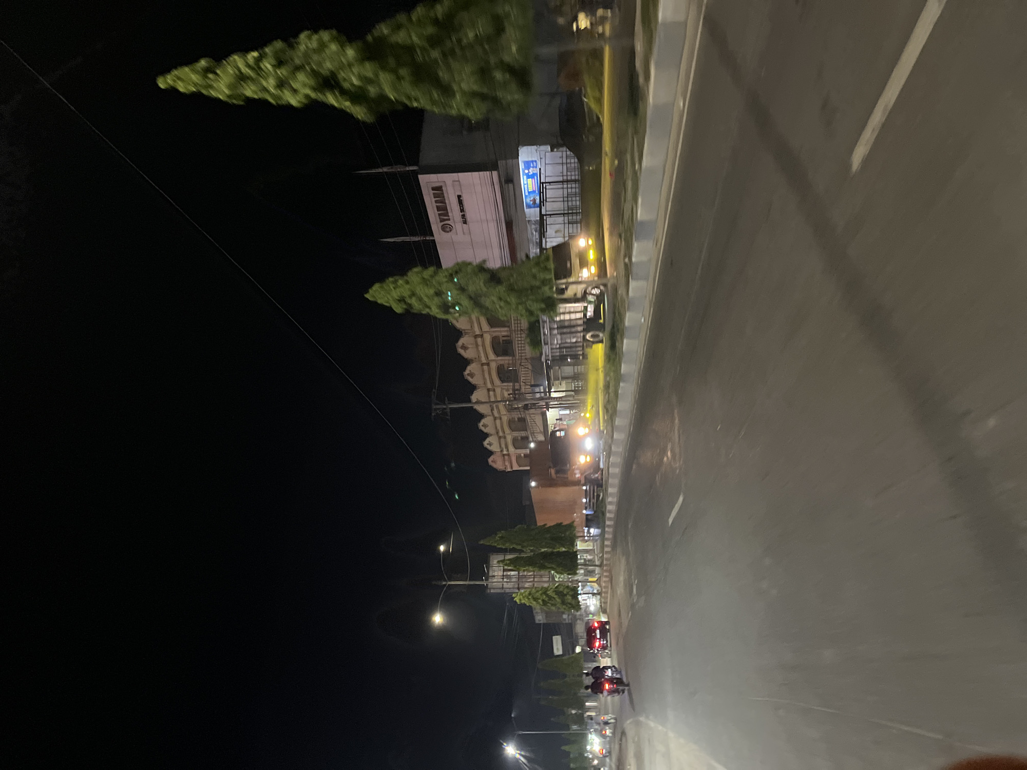 The image depicts a nighttime street scene in Bundar, Indonesia. The perspective is from a vehicle, likely a motorcycle, suggesting the photographer is a passenger or rider. The road is paved and appears to be a multi-lane thoroughfare, with a distinct dividing line. Streetlights illuminate the scene, casting a warm glow on the pavement and nearby structures.
On the left side of the road, several buildings are visible. One prominent building has a sign that reads "YAMAHA" in large, clear lettering, indicating a dealership or service center. Adjacent to this is another building with a sign in Indonesian script. Further down the street, another building with ornate architectural details, possibly a historical structure or a decorative facade, is lit up.
Several vehicles are present. The photographer's vehicle is moving, and other motorcycles and possibly cars are on the road ahead. The headlights of these vehicles are visible, cutting through the darkness. There are also streetlights and trees lining the side of the road, with some trees appearing to be ornamental.
The overall impression is that of a busy street at night. The lighting suggests it is well-lit for nighttime activity. The presence of multiple vehicles and illuminated businesses points to a commercial or residential area that is active even after dark. The image captures the typical urban nighttime atmosphere of a location like Bundar, Indonesia, with a blend of modern commerce and architectural elements.