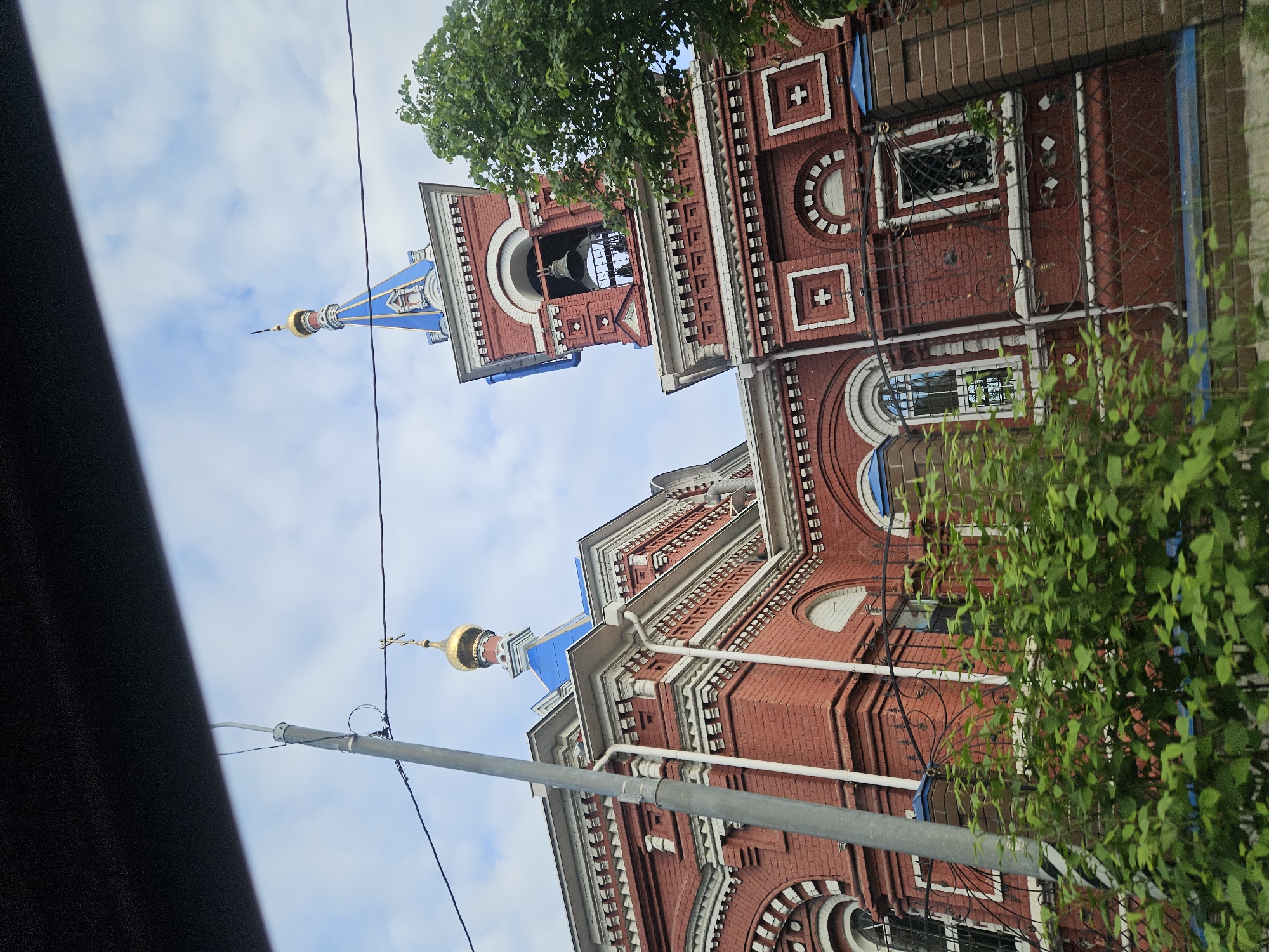 Here is a description of the image:

The image depicts the upward view of a red brick building with white accents and a blue roof, presumably a church. The structure features decorative elements such as crosses, circular windows, and arched openings. A bell tower rises from the main building. Parts of trees, including foliage, are visible in the foreground. Utility poles and wires cross the sky in front of the building. The image was taken in the city of Осеченки, Russia.