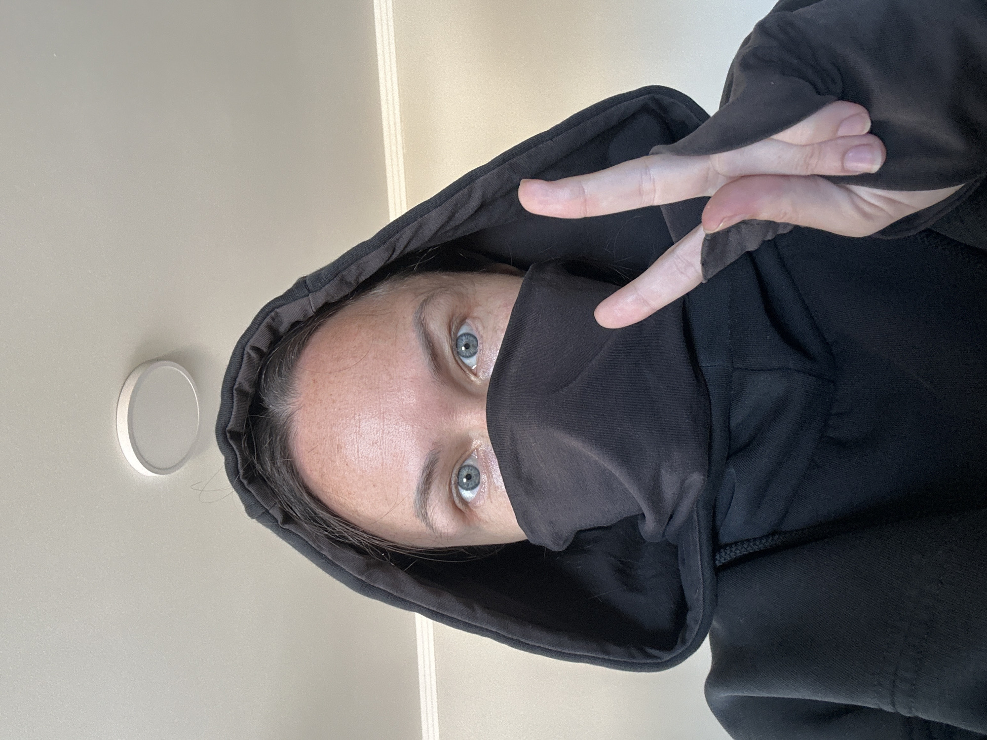 A close-up, low-angle shot depicts a person wearing a black hooded sweatshirt with the hood pulled up and partially covering their face. A section of the hoodie is pulled over their nose and mouth, obscuring the lower part of their face. The person's eyes, which are blue, are visible. The person's hand is raised, forming a peace sign with two fingers extended. The background appears to be an off-white or light beige wall, with a circular light fixture visible in the upper-left corner of the frame. The location is not discernible from the image alone, so the city of Florianópolis, Brazil, cannot be confirmed.