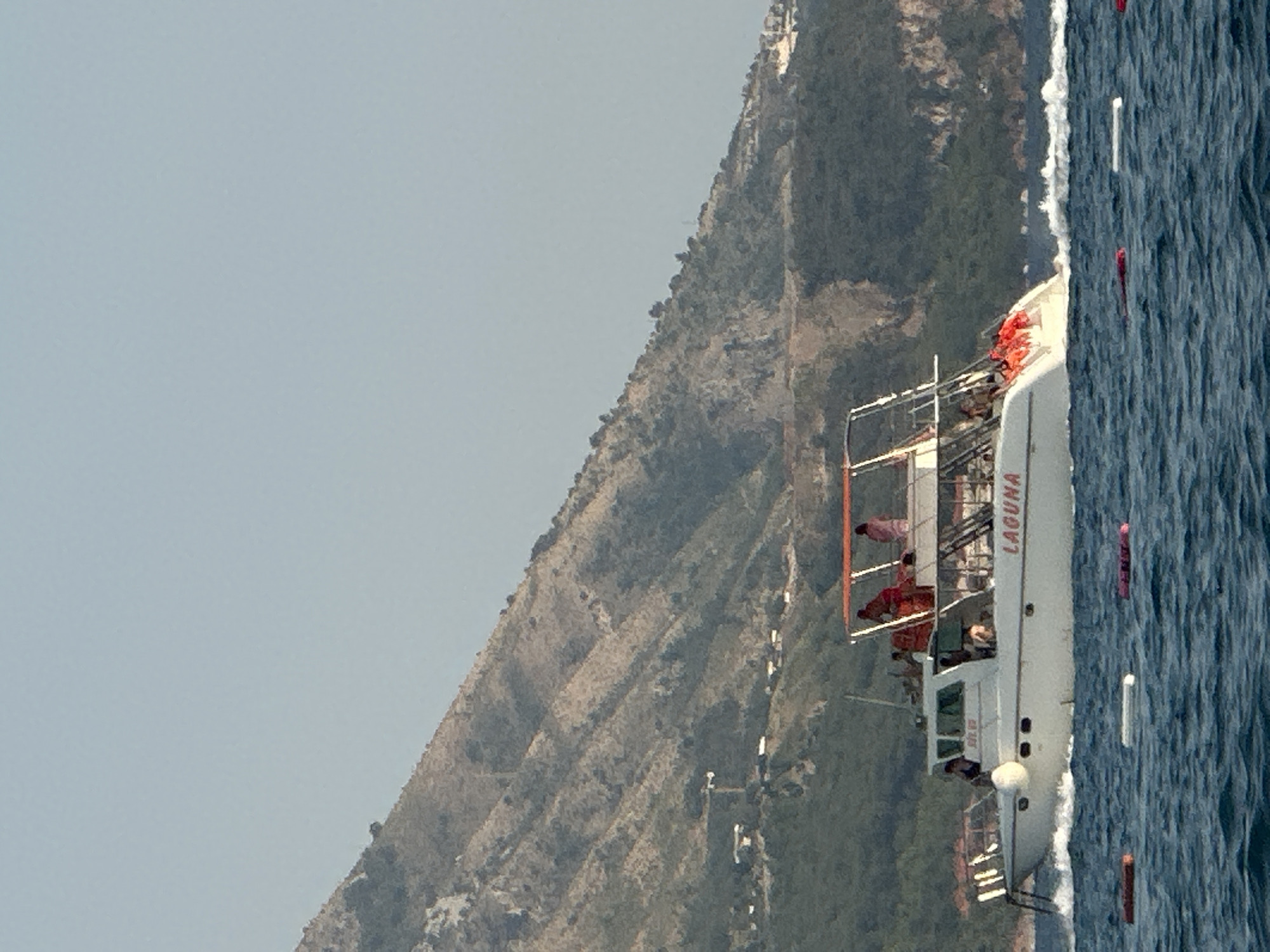 A white motor yacht, named "LAGUNA", travels across a body of water, leaving a white wake behind it. The yacht has multiple decks and an orange canopy structure. Several people, some wearing orange life vests, are visible on the yacht. In the foreground, several buoys float on the water. In the background, a steep, rocky mountain slope covered in vegetation rises. The sky is clear and pale blue. This scene is likely located near Boreti, Montenegro, given the characteristic mountainous coastline.
