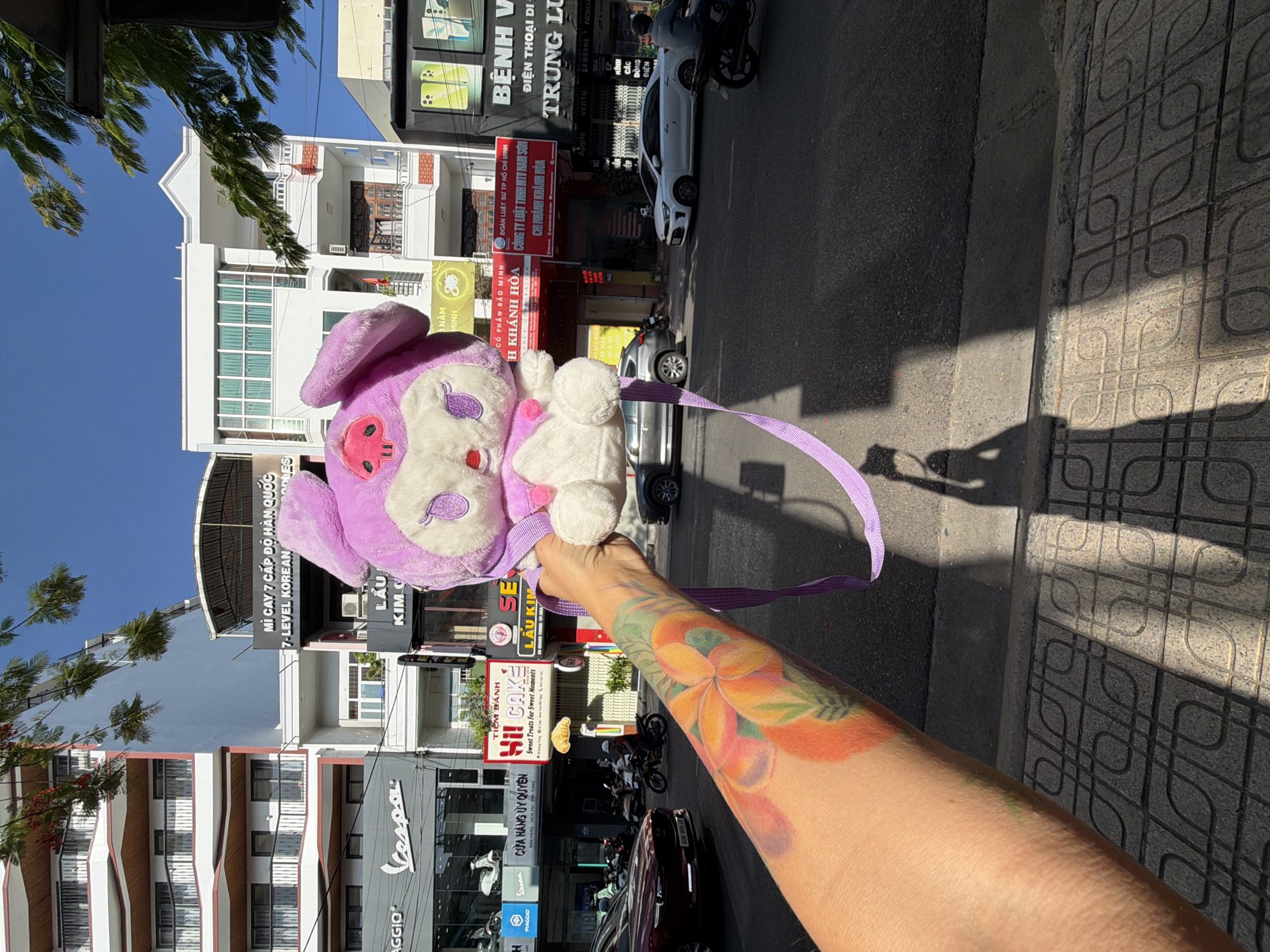 A first-person perspective shot captures a plush purple bunny backpack held up by an arm with a vibrant floral tattoo, set against a backdrop of a city street in Nha Trang, Vietnam. The street is lined with multi-story buildings, including a white building with a peaked roof and a building with balconies. Businesses visible on the street level include "Vespa," "Hi! CAKE," "LUU KIM," "SE LÀU KIM," and "BỆNH VIỆN ĐIỆN THOẠI." Various vehicles, including cars and motorcycles, are present on the street. The sky is clear and blue, and sunlight casts shadows on the pavement.