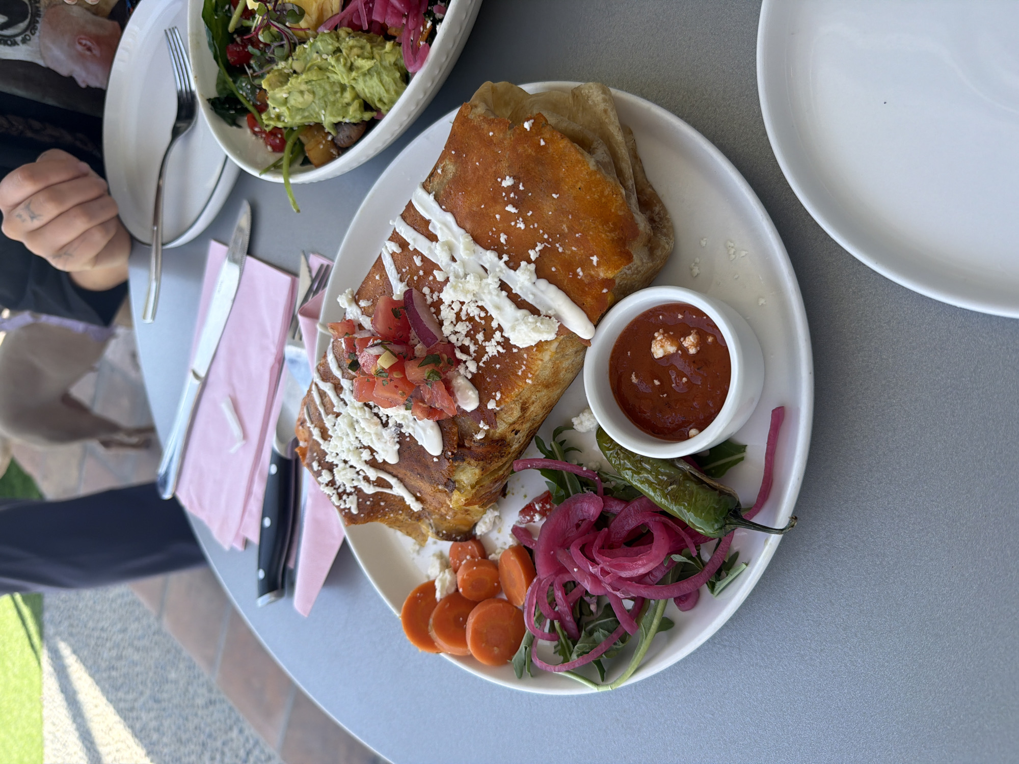An overhead shot captures a vibrant meal set on a light grey table, likely outdoors, given the visible tiled patio and greenery in the background. The primary focus is a large, golden-brown dish, possibly a fried burrito or chimichanga, topped with a drizzle of white cream or sour cream, crumbled white cheese (likely cotija), and fresh pico de gallo (diced tomatoes and red onions).
Accompanying the main dish on the white plate are several side items: a heap of thinly sliced pickled red onions on a bed of arugula or similar greens, several rounds of sliced cooked carrots, and a whole roasted green chili pepper. A small white ramekin holds a rich red salsa, possibly a salsa roja, flecked with white bits of cheese or herbs.
In the upper left quadrant, a person's left arm and hand are visible, holding a silver fork. Tattoos are visible on the fingers and hand. Further back and out of focus, part of another person's head and shoulder are seen, along with a white bowl containing a fresh salad topped with bright green guacamole, red pickled onions, and other vegetables, garnished with sesame seeds. An empty white dinner plate rests on the right side of the table. Cutlery, including a fork and two knives, is placed on pink napkins to the left of the main dish. The scene suggests a daytime meal, possibly brunch or lunch, in a relaxed outdoor dining setting. No discernible text is visible in the image.