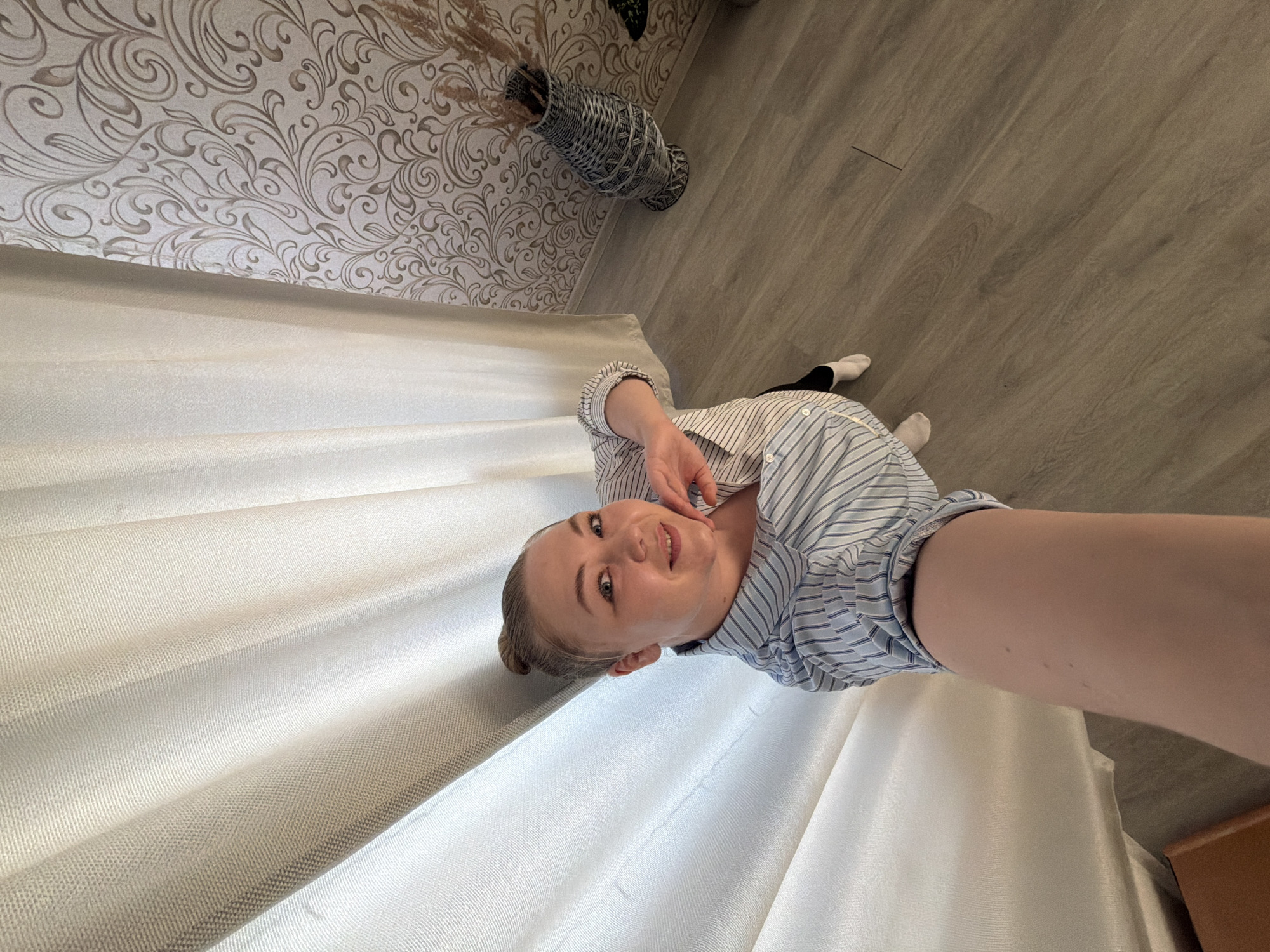 The image depicts a woman in a striped, button-down shirt, seemingly taking a selfie indoors. Her hand is near her face, and she is smiling. She is positioned near a white curtain in a room with patterned wallpaper and a grey, wood-like floor. A vase with dried plants is visible near the wallpaper. There is no evidence to suggest the location is in the city of Noviy, Russia.