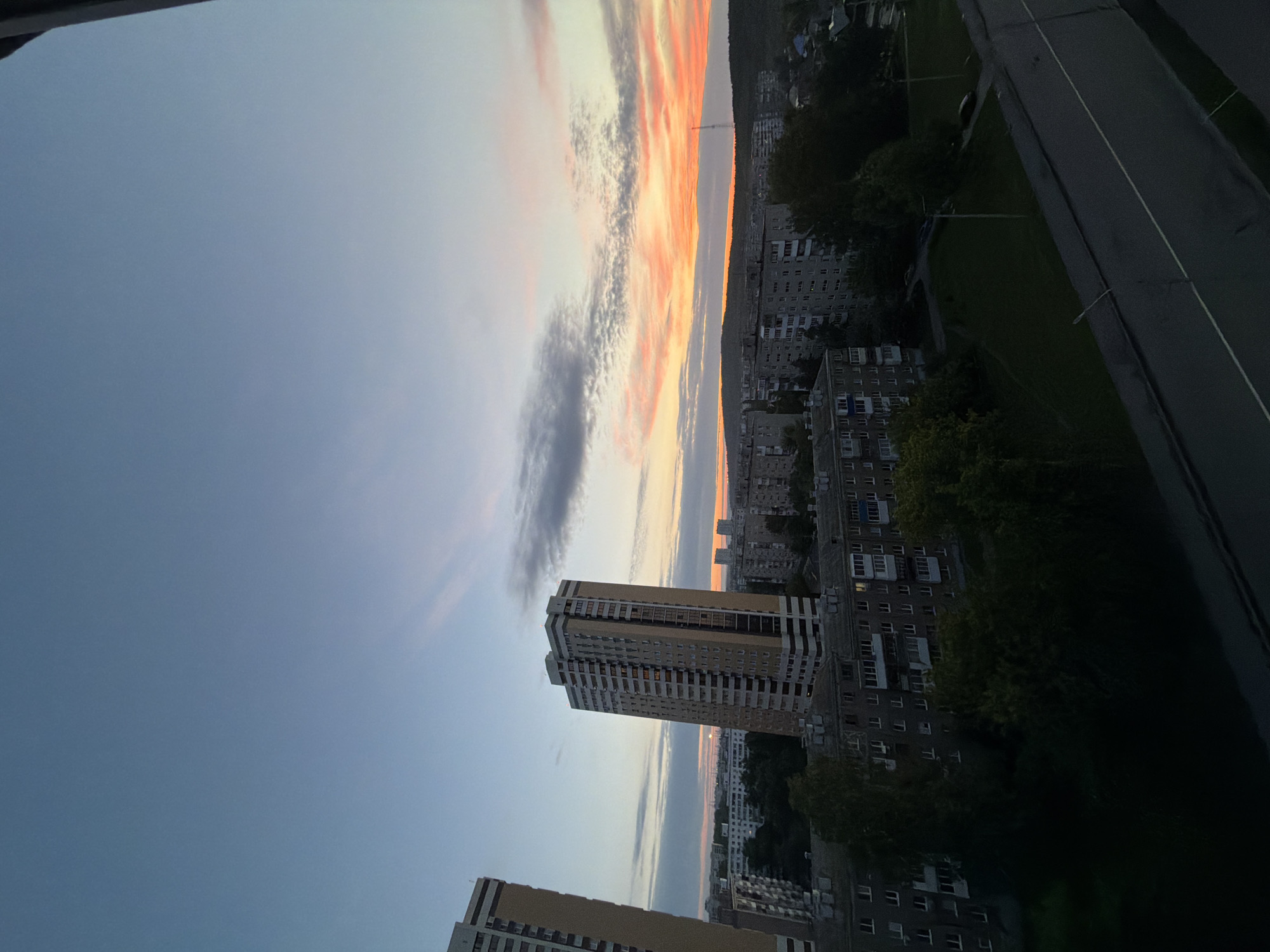 An elevated outdoor wide shot shows a city at dusk. The sky is a gradient of blue and orange, with streaks of clouds illuminated by the setting sun. In the foreground, a dark, angled surface, possibly a roof or balcony, dominates the bottom of the frame. Beyond this, a road with a white line marking the lane can be seen. Further into the scene, there are residential buildings of varying heights. Two tall, slender buildings with light-colored facades and dark window frames stand out, one on the left and a more central one. Between these, and extending towards the horizon, are lower-rise apartment buildings, many appearing to be made of concrete. Dense green trees are interspersed among the buildings, some obscuring the lower levels. In the distance, the city stretches towards a dark, hilly horizon. The overall lighting suggests either early morning or late evening.