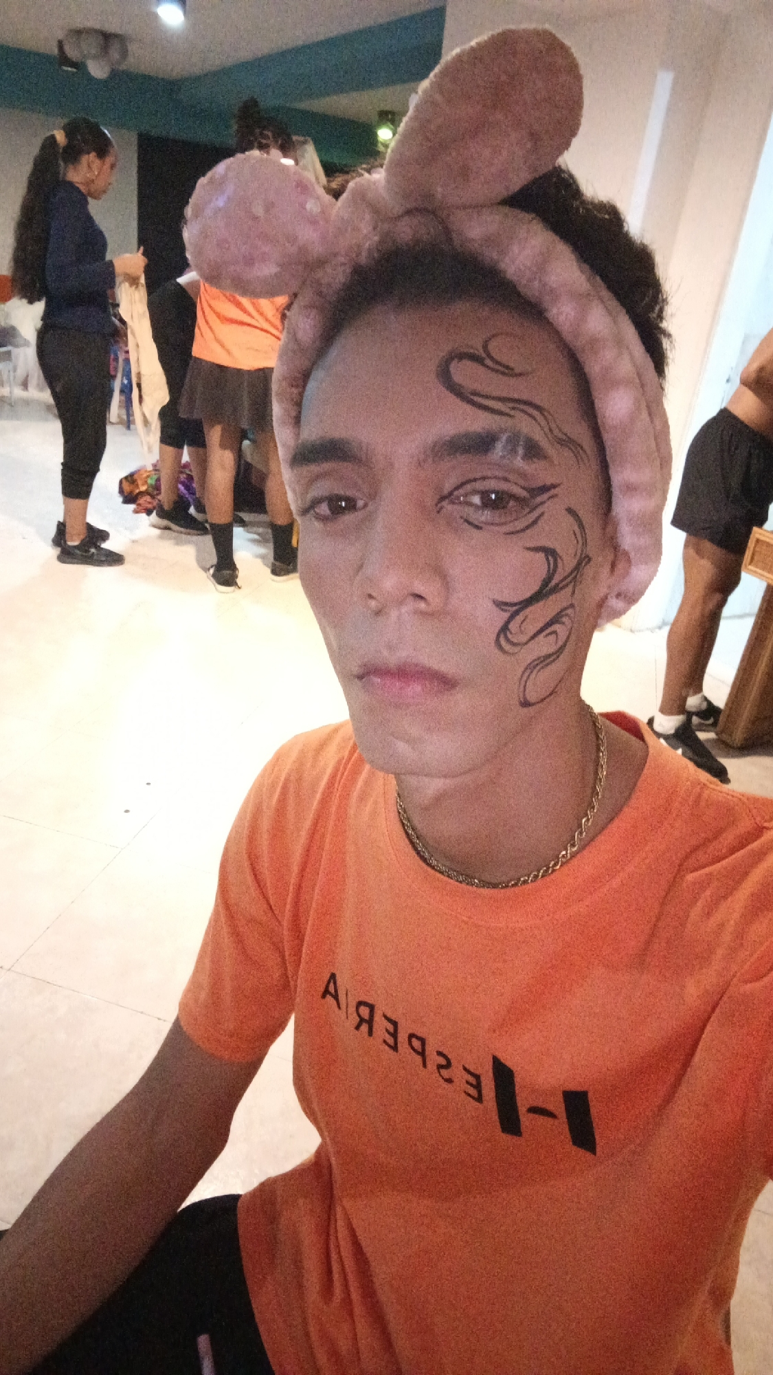 The image is a selfie taken indoors. A person wearing a peach-colored headband with bunny ears and black face paint swirls around their left eye is centered in the frame. They have an orange T-shirt with a reversed black logo and a gold chain around their neck.
In the background, several people are milling about. One woman is wearing a dark long-sleeved shirt and dark pants, and another is wearing an orange shirt and dark skirt. Additionally, a small section of another person wearing black shorts is visible. The lighting is bright and somewhat uneven.
The city of Playa El Agua, Venezuela is not discernible from the contents of the image.