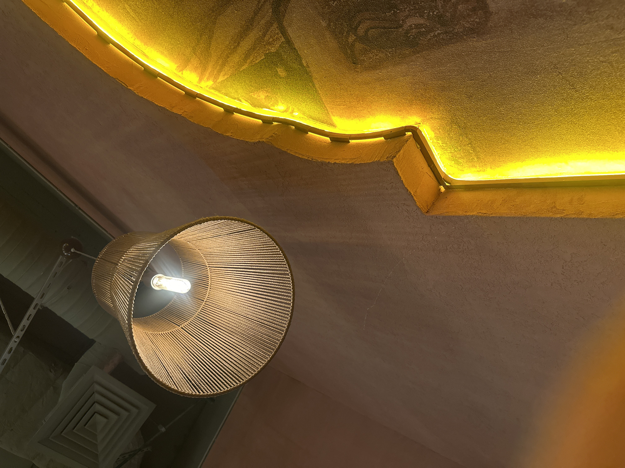 A low-angle shot depicts an interior setting, likely in Kazan, Russia, showcasing elements of design and architecture. A woven, cylindrical lampshade with a visible illuminated bulb hangs from the ceiling on the left, accompanied by the supporting structure. The ceiling is textured and appears to be painted in a muted tone. A linear, bright yellow neon light fixture is embedded within a recess in the ceiling above, illuminating the space. The image also captures a portion of an air vent on the lower left.