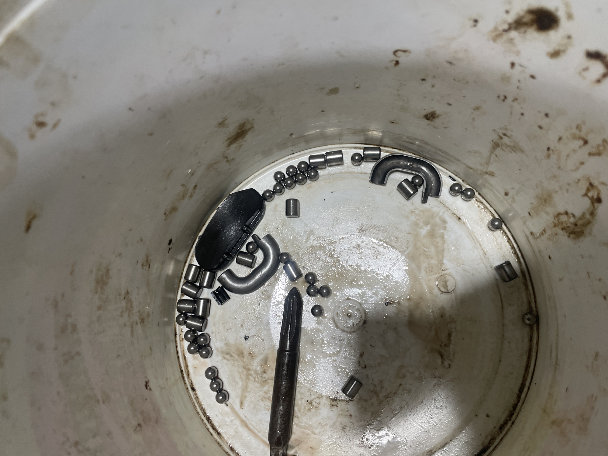 The image shows the interior of a dirty, white plastic bucket. Inside are various metallic components including ball bearings, cylindrical rollers, and curved metal pieces, along with a black plastic object and a metal tool resembling a punch or awl. The bucket's interior is stained with a dark substance, possibly grease or oil. No people are visible in the image. The location is not discernible from the visual information.