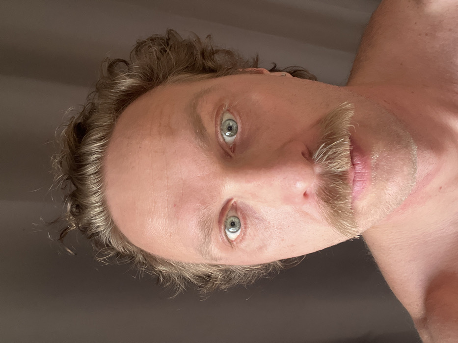 The image depicts a close-up, head-and-shoulders portrait of an adult male. He has light skin, light brown curly hair, and blue-green eyes. A full blonde mustache is prominent above his lips, and stubble is visible on his chin and jawline. A small silver stud earring is located on his right earlobe. His shoulders and upper chest are bare, with a faint reddish mark present on the upper left chest. The male subject gazes directly into the camera with a neutral, wide-eyed expression. The background is an out-of-focus, solid dark grey or taupe surface, consistent with an indoor setting like a wall or curtain. No discernible features or objects are present within the image to identify a specific location within the city of Saint Petersburg, Russia.