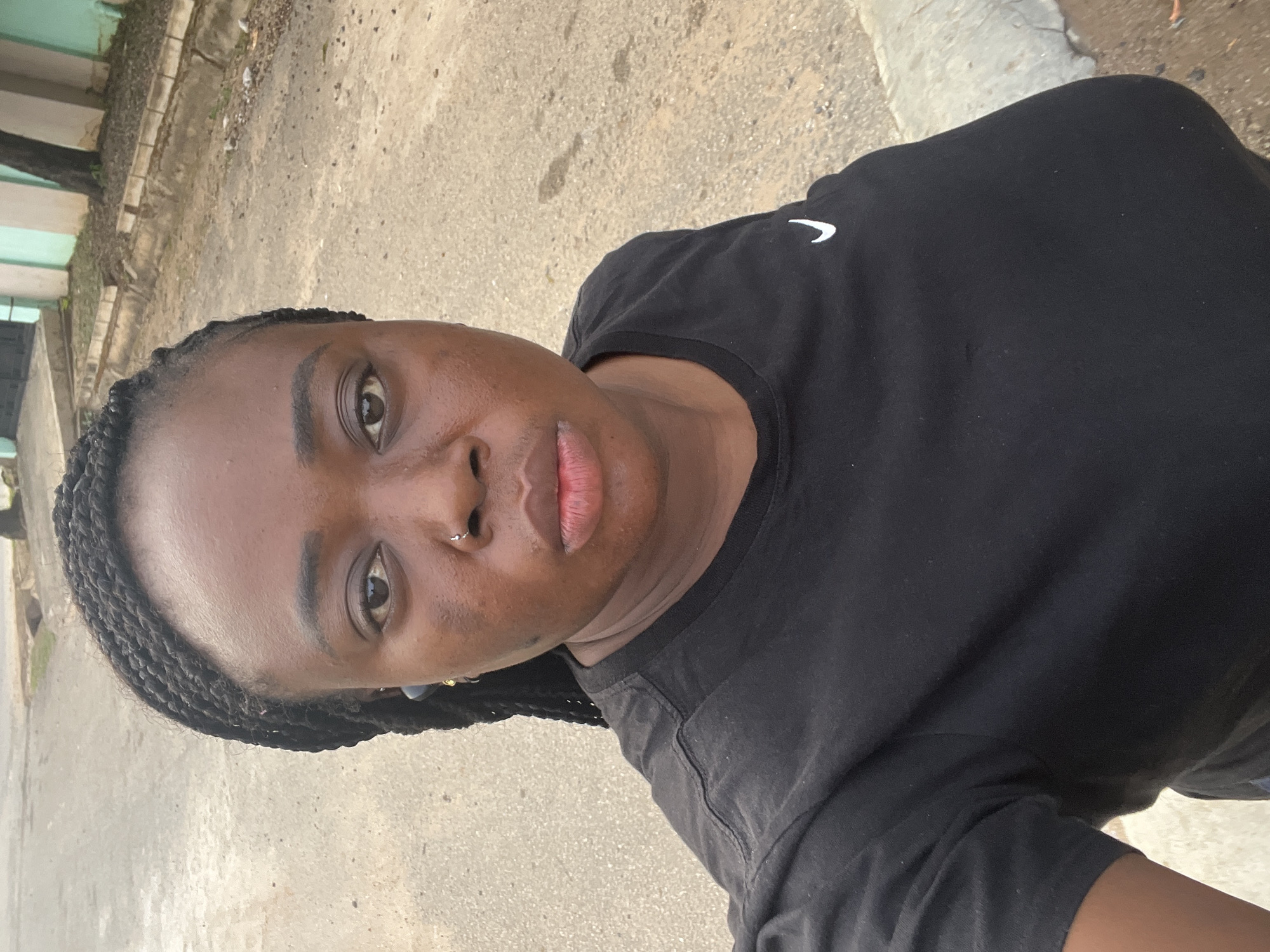 The image is a vertical full-frame selfie of a dark-skinned individual, appearing to be a young adult, looking directly into the camera. The person is captured from the chest up, with their head slightly tilted. They have dark, neatly braided hair styled away from their face, dark eyes, and a small, silver hoop nose ring on their right nostril. Their lips show a subtle reddish tint. The individual is wearing a black t-shirt featuring a white Nike swoosh logo on the upper left chest.
The background indicates an outdoor, urban or suburban setting, photographed during daytime with diffused natural lighting. Behind the person, a textured, light brown/greyish ground surface, possibly a dirt or concrete path, is visible. To the left, a portion of a light turquoise-green building or wall can be seen, with a dark tree trunk partially obscuring it. A raised concrete curb or pathway edge runs diagonally through the mid-ground on the left. The individual's expression is neutral and calm. The overall scene suggests a casual, everyday moment in a typical outdoor environment in Abuja, Nigeria.