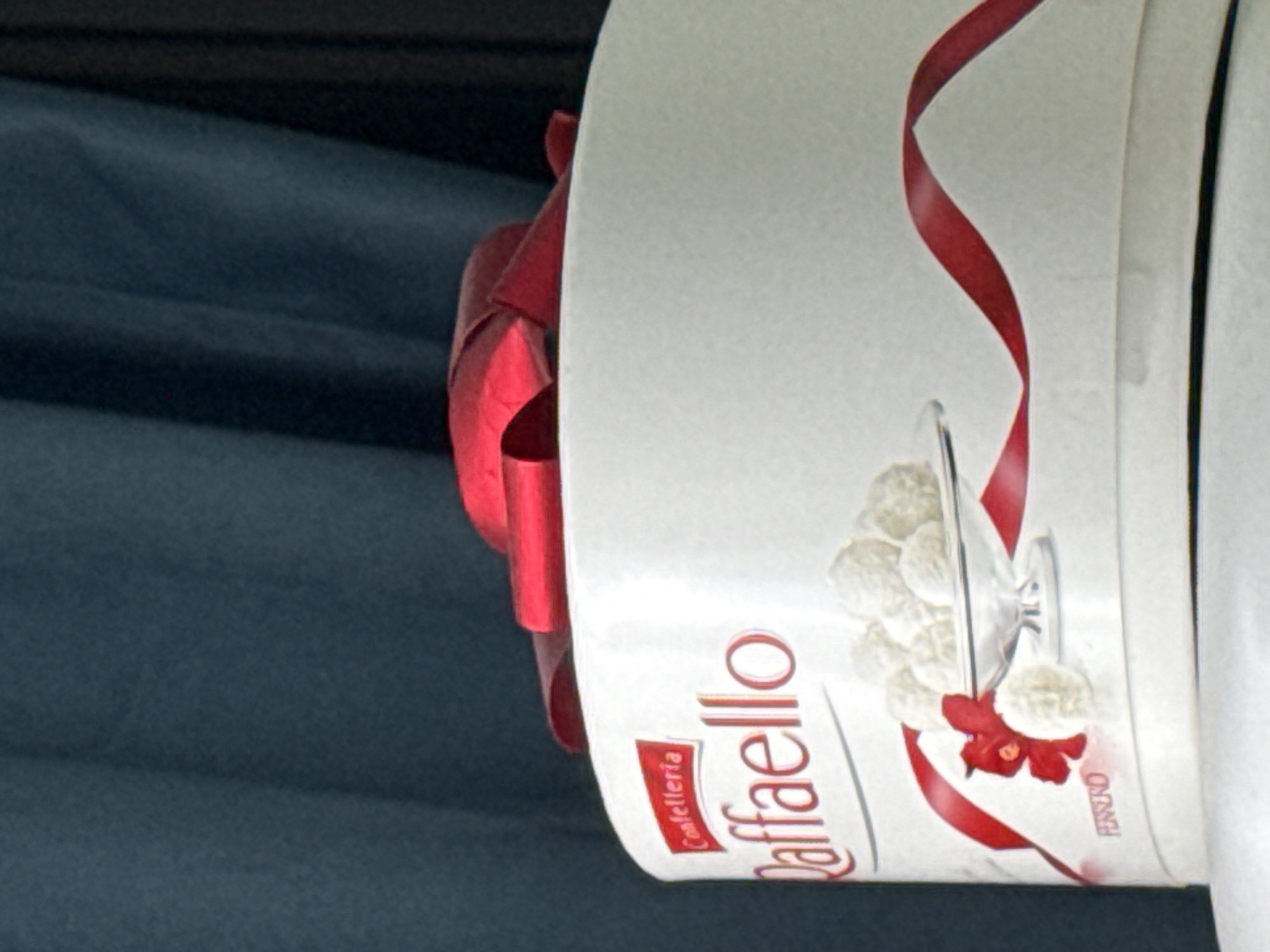 The image shows a close-up of a round white box of RaffaHELLO chocolates. The box has a red ribbon tied around the lid, and the brand name "Raffaello" and "Confetteria" are printed on the side in red lettering. A small red bow is visible on the front of the box. Inside the box, there is a clear glass dish filled with white RaffaHELLO chocolates. Below the dish, the word "FERRERO" is printed in small purple letters. The background is a dark blue curtain. There are no people or animals in the image, and no indication of the location being in Elista, Russia.