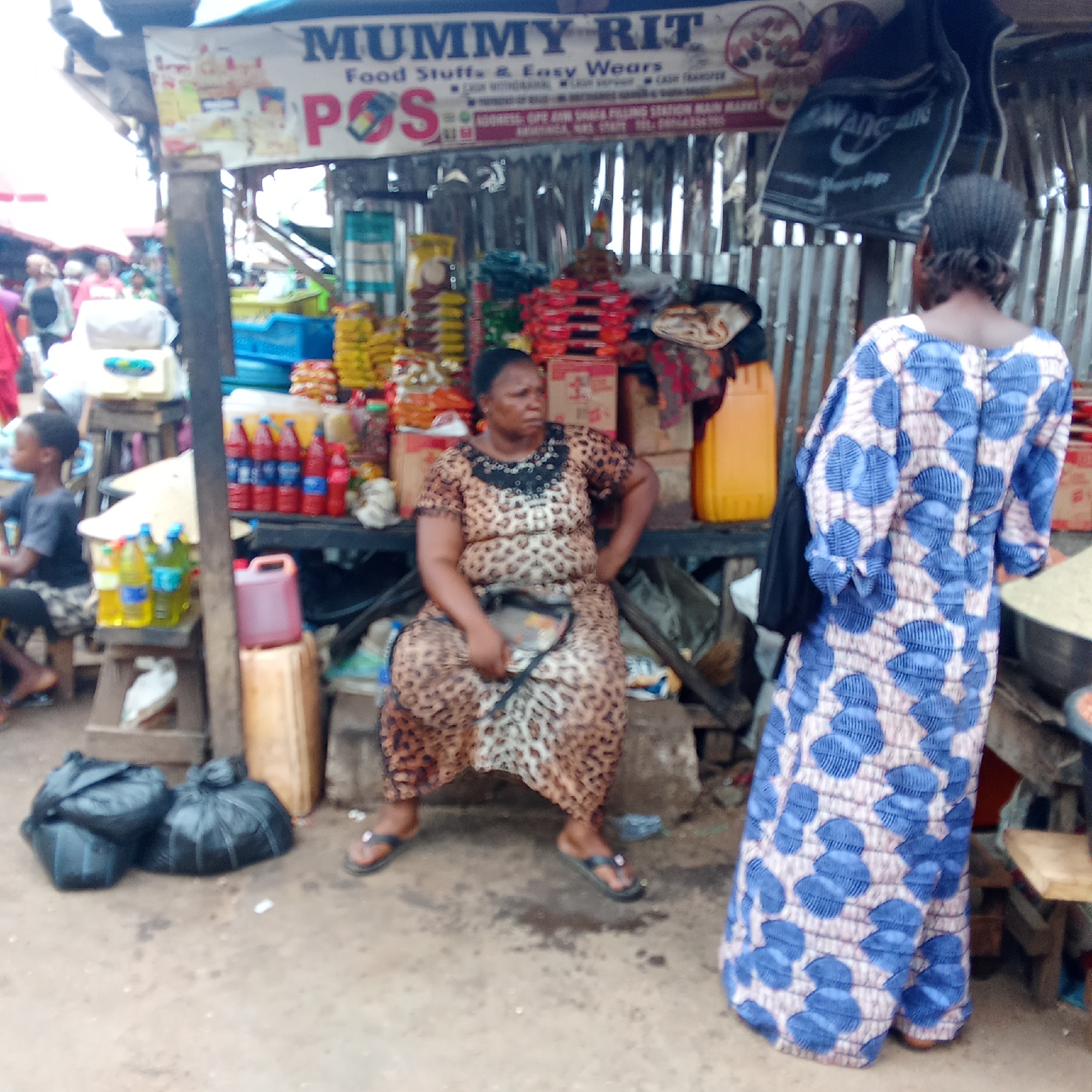 The image depicts a market scene in Akwanga, Nigeria. A woman wearing a leopard-print dress sits behind a stall filled with food items and other goods. The stall has a sign that reads "MUMMY RIT Food Stuffs & Easy Wears POS ADDRESS: OFE AWA SHAJA FILLING STATION MAIN MARKET". In front of the stall, there are two black bags on the ground. To the right, a woman wearing a blue and white patterned dress stands with her back to the camera. A young boy wearing a gray shirt sits to the left of the stall. The background shows a bustling market environment with other people and stalls visible.