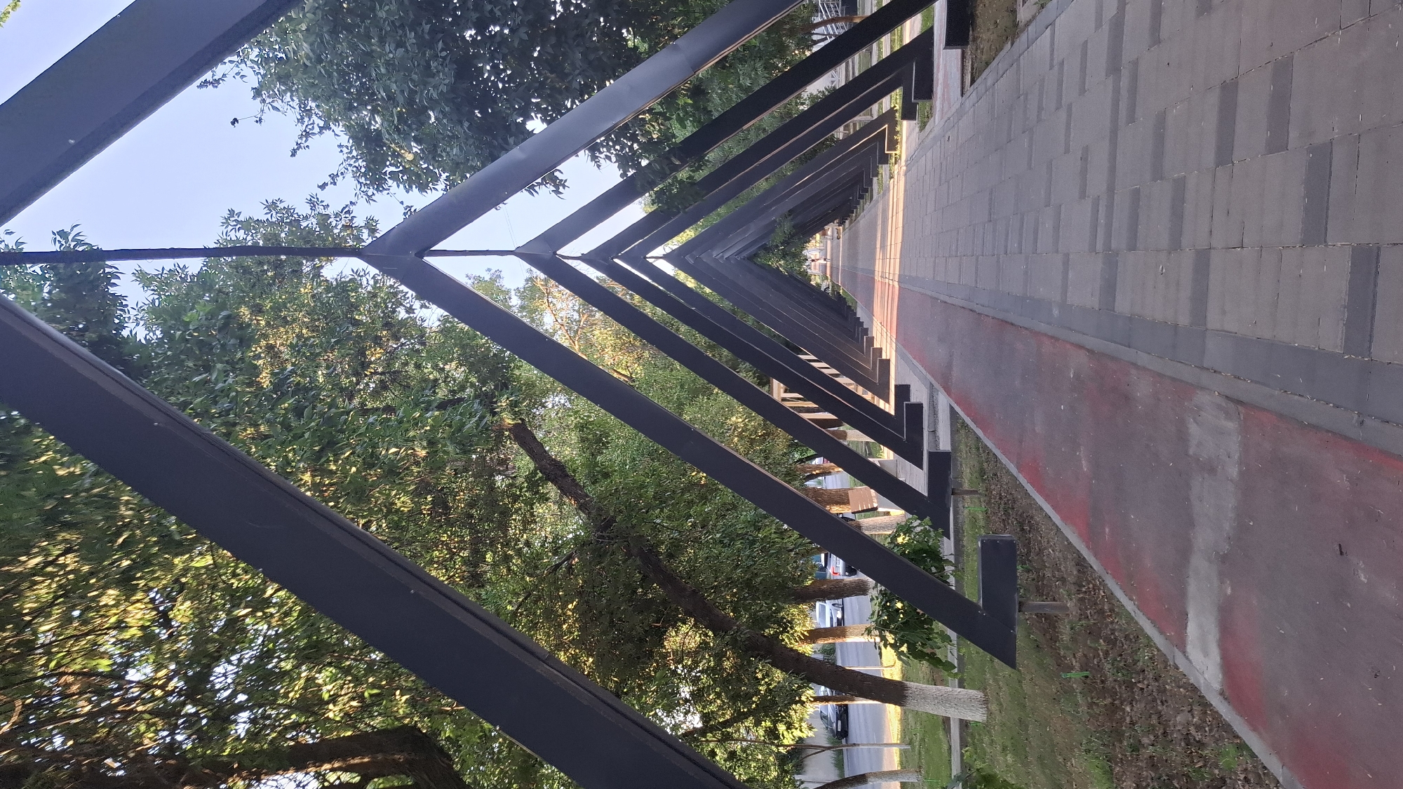 The image shows an outdoor pedestrian walkway in Shymkent, Kazakhstan. The walkway is lined on one side by a series of triangular, dark-colored metal structures forming an arched canopy. The walkway itself is paved with rectangular stone tiles. To the left of the tiled walkway is a red-colored path, possibly for cycling or running. Beyond this path is a grassy area with trees and white-trunked trees. In the background, cars are visible on a street, and more trees line the area. The sky is clear and blue. The perspective suggests the photo was taken from a low angle, looking down the length of the walkway.