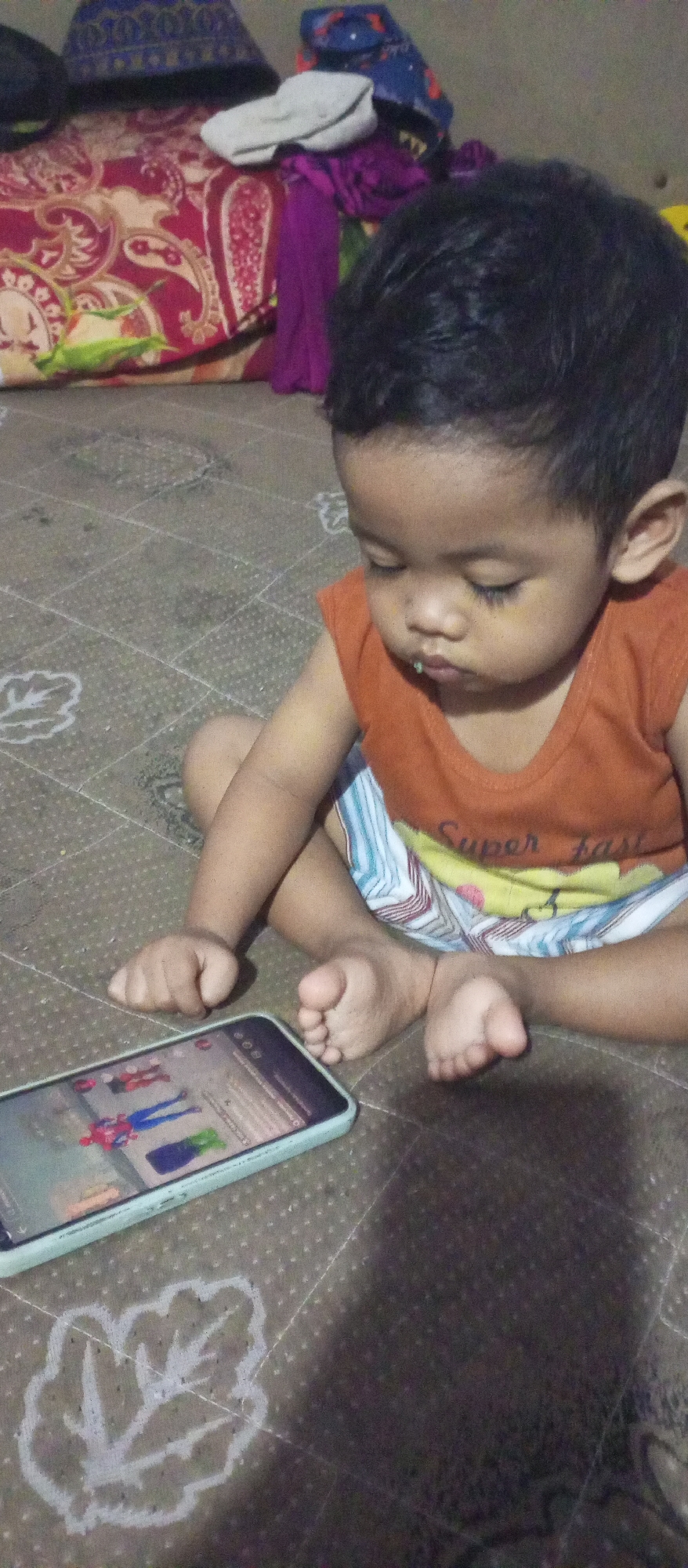 A young boy with dark hair and an orange sleeveless shirt is sitting cross-legged on a patterned floor. His eyes are closed, and he appears to be looking at a smartphone that is placed in front of him. A small amount of green food is visible on his upper lip. His legs are bent, and his feet are resting on the floor. The smartphone screen displays an image of what appears to be characters from a video game, possibly superheroes like Spider-Man. In the background, there are some folded fabrics, including a red and beige patterned cloth, a purple cloth, and a white cloth, as well as a dark blue object with a pattern. The setting seems to be an indoor environment, possibly a home. The image was taken in Rejoso Kidul, Indonesia.