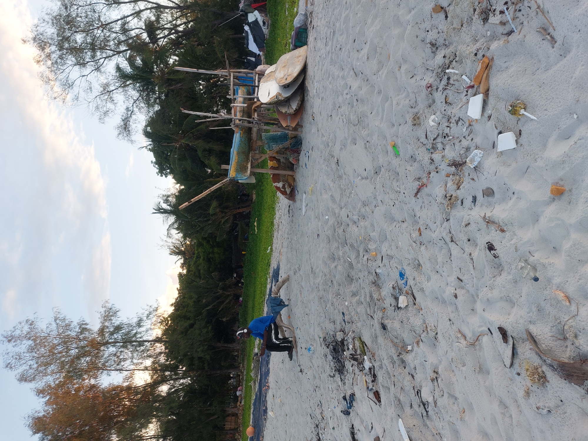 The image depicts a beach scene in Dar es-Salaam, Tanzania. A man in a blue shirt and dark pants is bending over, possibly collecting trash, on a sandy beach littered with debris. To the left is a dense area of green trees and foliage. In the background, there is a wooden structure with various objects attached, including what appears to be surfboards. The sky is visible in the upper left corner, showing some clouds. The beach is scattered with trash.