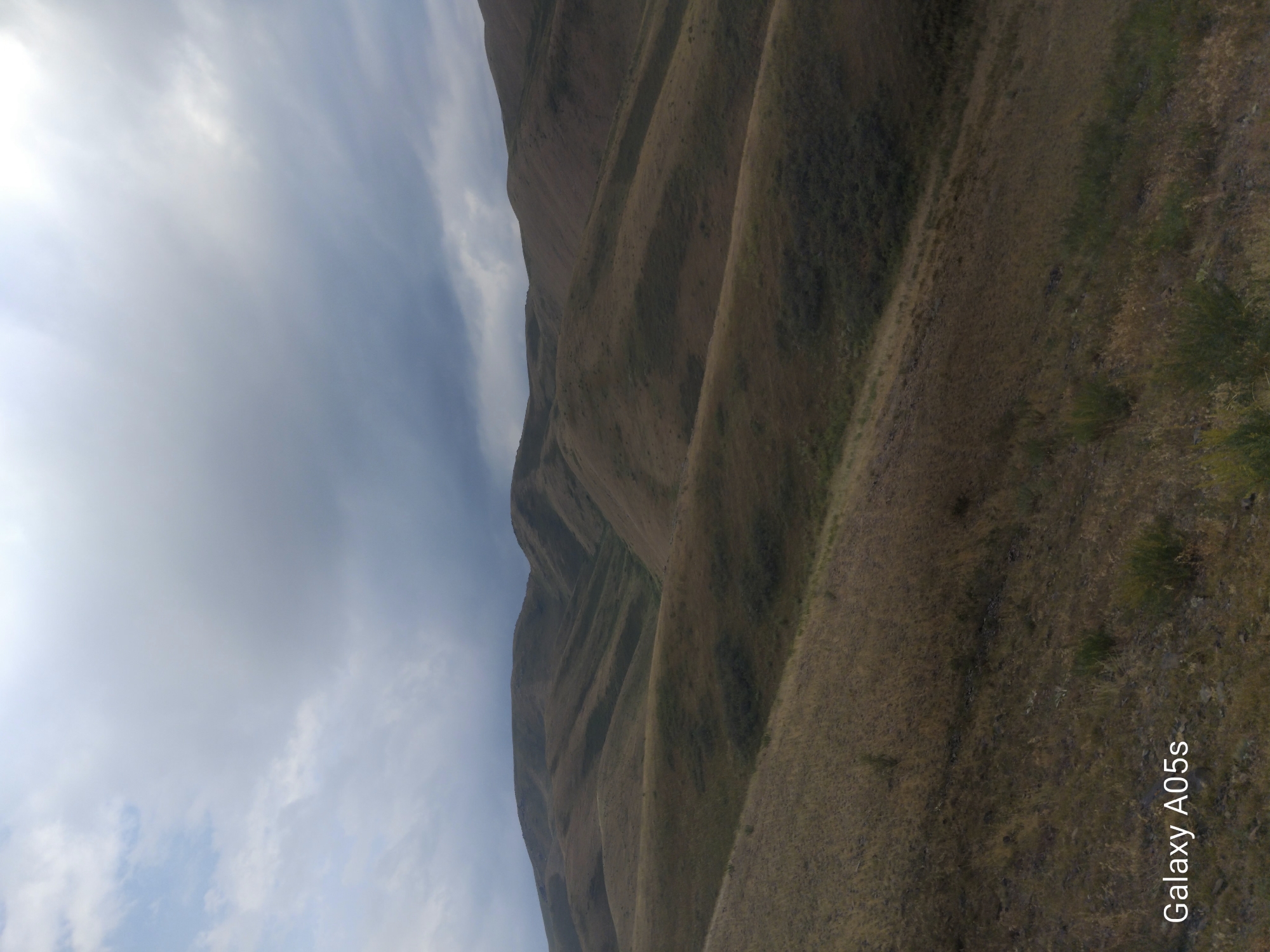 A landscape photograph captures a view of rolling hills under a cloudy sky. The foreground shows dry, brown grass and scattered green bushes on a sloped terrain. The middle ground and background are dominated by undulating hills covered in similar dry vegetation, with shadows defining their contours and indicating the direction of light. The sky is overcast with heavy gray clouds, obscuring the sun and creating a diffused light across the scene. The bottom left corner of the image has a watermark that reads "Galaxy A05s". The location is identified as Kopuro Bazar, Kyrgyzstan. There are no people or visible animals in the frame, and no specific actions or interactions are depicted. The scene is natural and undeveloped.