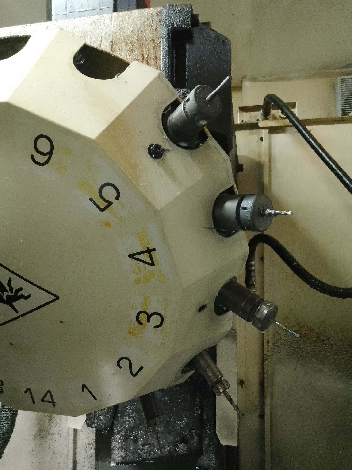 The image displays a close-up view of a multi-spindle rotary tool changer, a component commonly found on CNC machining centers. The tool changer is light beige with black numbers indicating different tool positions, ranging from 1 to 14, arranged in a circular pattern. Several tool holders, each containing a drill bit, are mounted in different positions on the changer. Some of the drill bits are visible, showing their sharpened tips.
The tool changer is attached to a larger, dark grey industrial machine. A black flexible hose, likely for coolant or air, extends from the machine towards the tool changer. The background consists of a wall and some metallic structures, suggesting an industrial or workshop environment. The surfaces of the machine and the surrounding area show signs of use, with some dirt and smudges.
There are no people or animals visible in the image. The scene is static and appears to be an interior shot of a manufacturing facility. The image is an objective depiction of industrial machinery. The context of Ciantra, Indonesia is not directly conveyed by visual elements within the image itself.