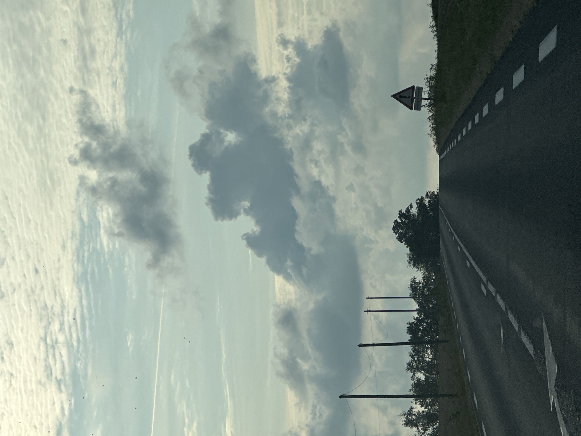 The image captures a view of a road leading uphill towards a horizon obscured by clouds and a hazy sky. Several dark poles with wires stretched between them are visible on the left side of the road, interspersed with dense green trees. On the right side of the road, a triangular warning sign with an exclamation mark is prominently displayed, situated on a grassy embankment. The road itself is paved and marked with white dashed lines indicating lane separation. The sky is filled with a mixture of textured, light-colored clouds and larger, darker, more dramatic cloud formations. There is a faint contrail visible in the upper left quadrant of the sky. No people or vehicles are present in the image. The overall impression is a somewhat overcast day on a rural road in Saint-Maclou-de-Folleville, France.