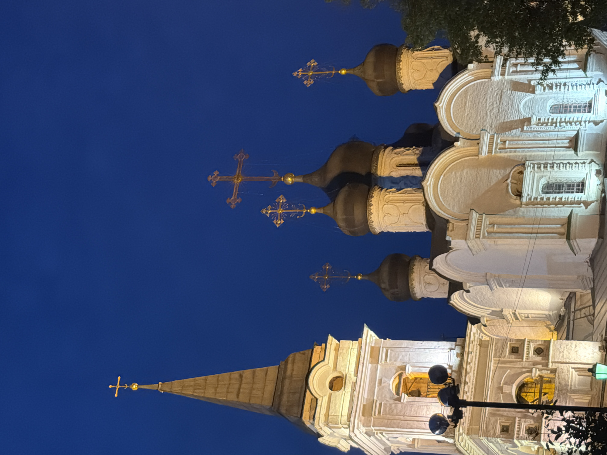 The image shows a dimly lit exterior shot of the Church of the Transfiguration in Tverskaya Zastava Square, Moscow, Russia, at dusk or night. The church features several domes topped with Orthodox crosses, illuminated by artificial lighting. A bell tower with a spire is visible on the left side of the frame, also lit. The sky is a deep blue, indicating twilight or nighttime. There are no people visible in the image. The overall scene is serene and architectural.
