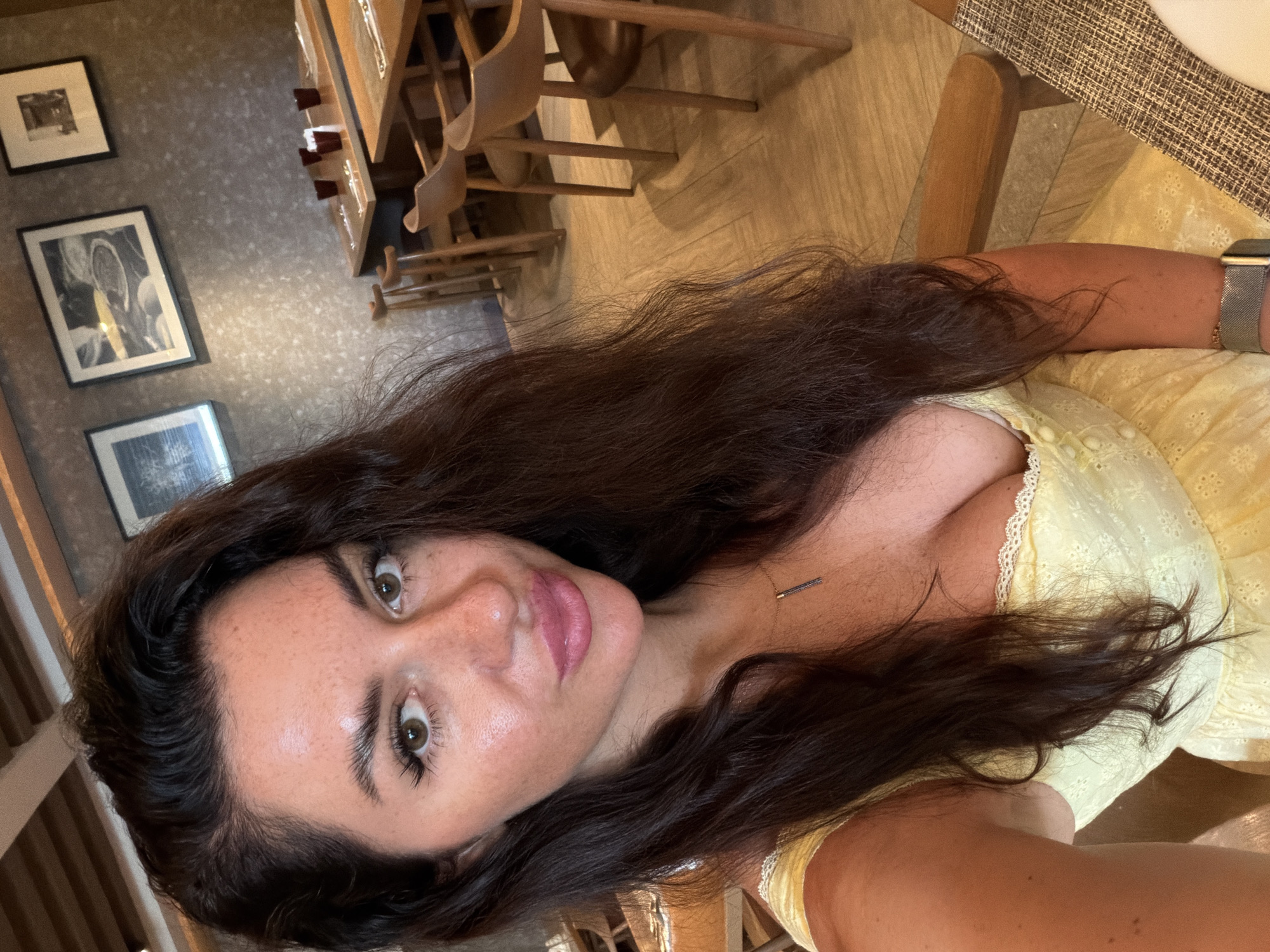 A woman with long, dark, wavy hair and light brown eyes takes a selfie in a dimly lit restaurant. She is wearing a yellow sundress with white lace trim and a silver necklace. Her hair is falling over her face, partially obscuring her left eye and ear. In the background, several wooden tables and chairs are set up for dining, with framed artwork on the wall and wooden beams on the ceiling. The location is the city of Bang Muang, Thailand.