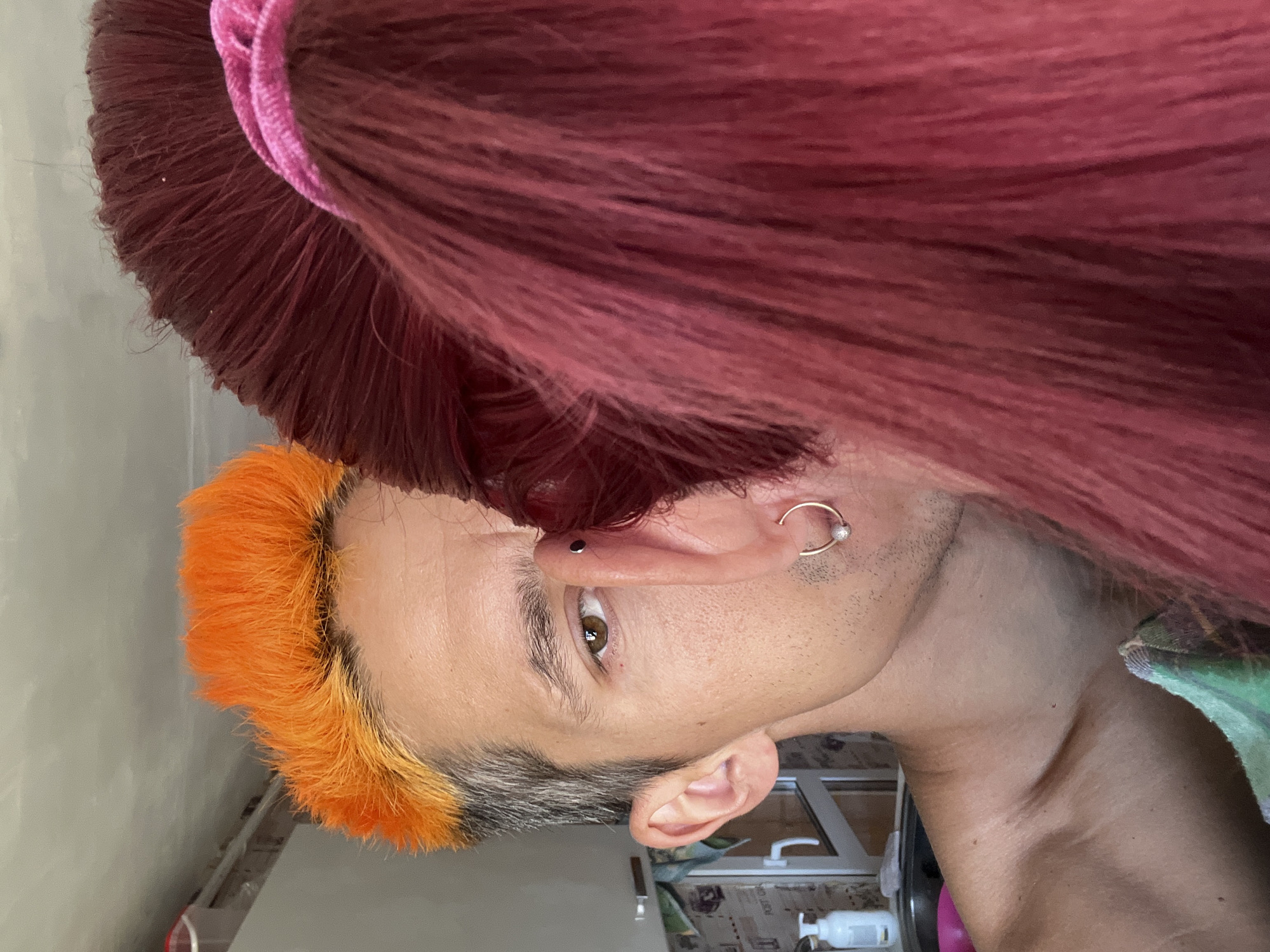 An indoor close-up depicts two individuals in the city of Chaikovsky, Russia. The individual on the left, identified as male, has short, spiky, bright orange hair on top that transitions to natural grey/darker hair on the sides. His right eye, brown in color, is visible along with part of his forehead, eyebrow, nose, cheek, and bare right ear, which has no visible piercings. Fine stubble is present on his jawline and lower cheek. His bare right shoulder and collarbone are visible in the lower left. A small red mark is present below his right eye. The second individual, positioned on the right and mostly out of frame, has long, dark red/maroon hair pulled back, secured by a pink fabric scrunchie. Their left ear is partially visible, featuring a black stud piercing in the upper cartilage (helix) and a gold hoop earring with a small white pearl or bead in the lobe. The second individual's hair and ear obscure the left side of the first individual's face and body. In the background on the left, a white pump bottle rests on a countertop beside a visible sink rim and white cabinet doors with silver handles. A green and white checkered fabric is draped in the lower left foreground. The lighting is bright and even.