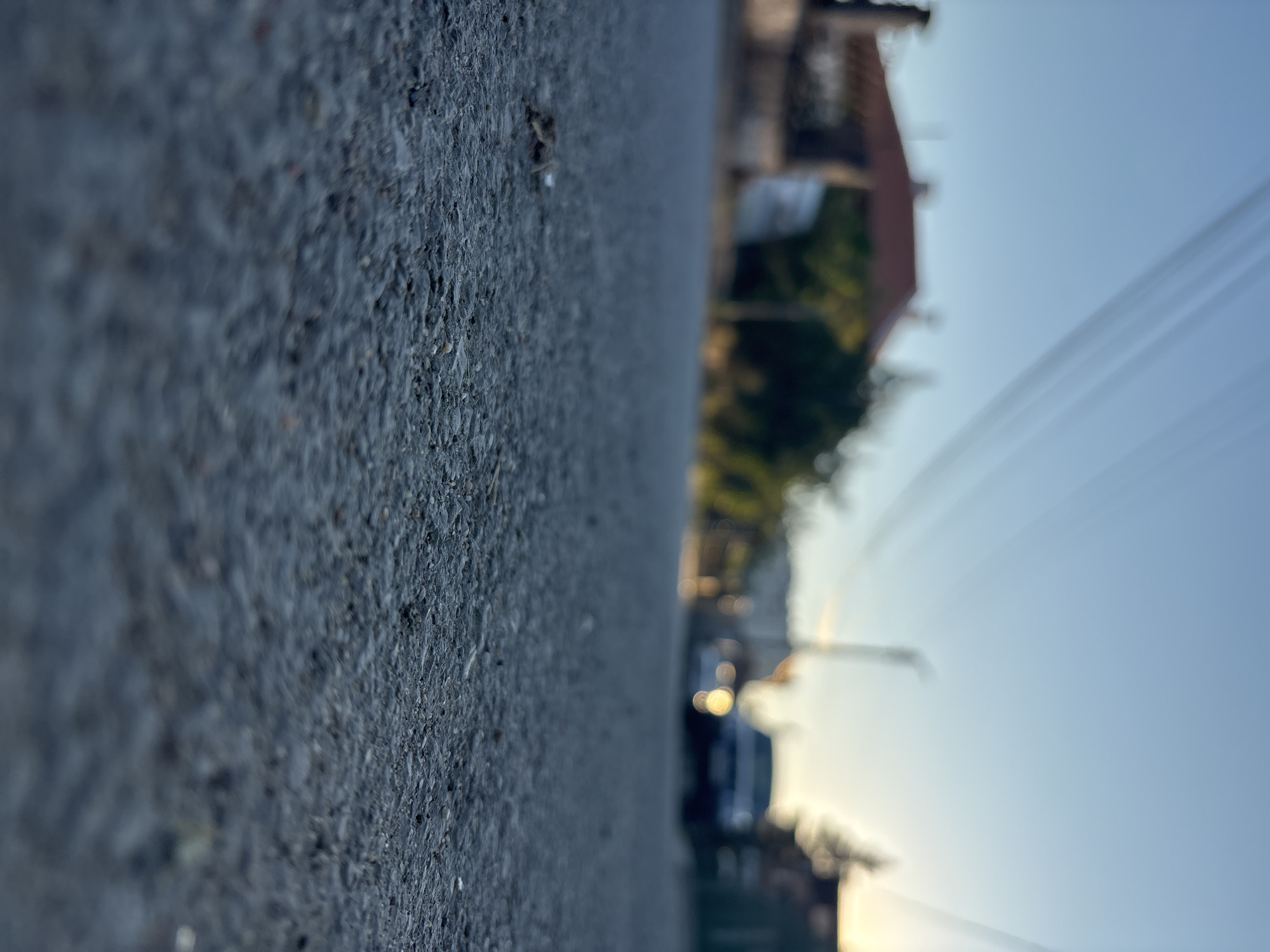The image shows a street in Lom, Bulgaria, taken from a low angle, focusing on the asphalt surface. Buildings are visible in the background, including a house with a red roof. Power lines stretch across the sky. A car is parked on the street, blurred in the distance. The sky is a light blue, possibly indicating dawn or dusk.