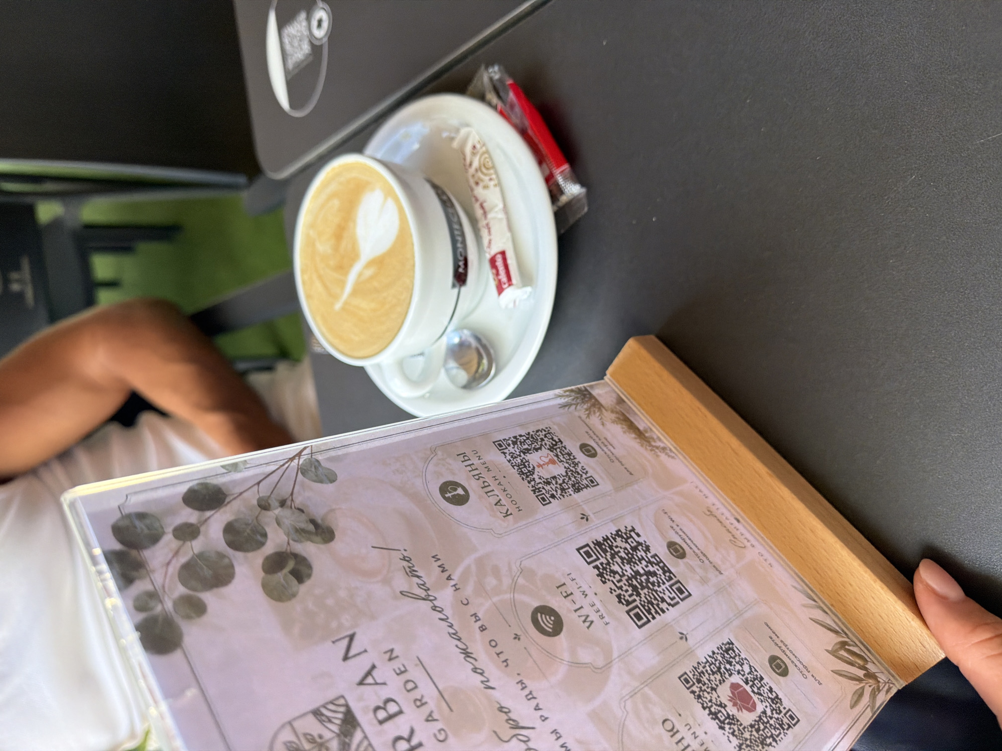 This is a medium shot, taken from a high angle, of a person's hand resting on a menu at a table. The menu is made of clear plastic and is framed by a light-colored wood. The menu displays several QR codes and text in both Russian and English. Visible text includes "URBAN GARDEN," "ДО ПОЖАЛОВАТЬ! МЫ РАДЫ, ЧТО ВЫ С НАМИ" (meaning "Welcome! We are glad that you are with us"), "WI-FI FREE WI-FI," and "КАЛЬЯНЫ HOOKAH MENU."
To the left of the menu, a cup of coffee with latte art is visible, placed on a saucer. A spoon and a sugar packet are also on the saucer. The coffee cup has the word "MONTECELE" printed on it. In the background, a person's arm and leg, wearing a white garment, are visible. The setting appears to be an outdoor cafe, with hints of greenery in the background, possibly a garden area. The overall lighting suggests it is daytime, likely sunny. The scene evokes a relaxed atmosphere, perhaps during a leisurely break. The presence of the menu with QR codes suggests a modern dining establishment. The mention of "La Vila Joiosa, Spain" in the prompt implies the location is a coastal town in Spain.