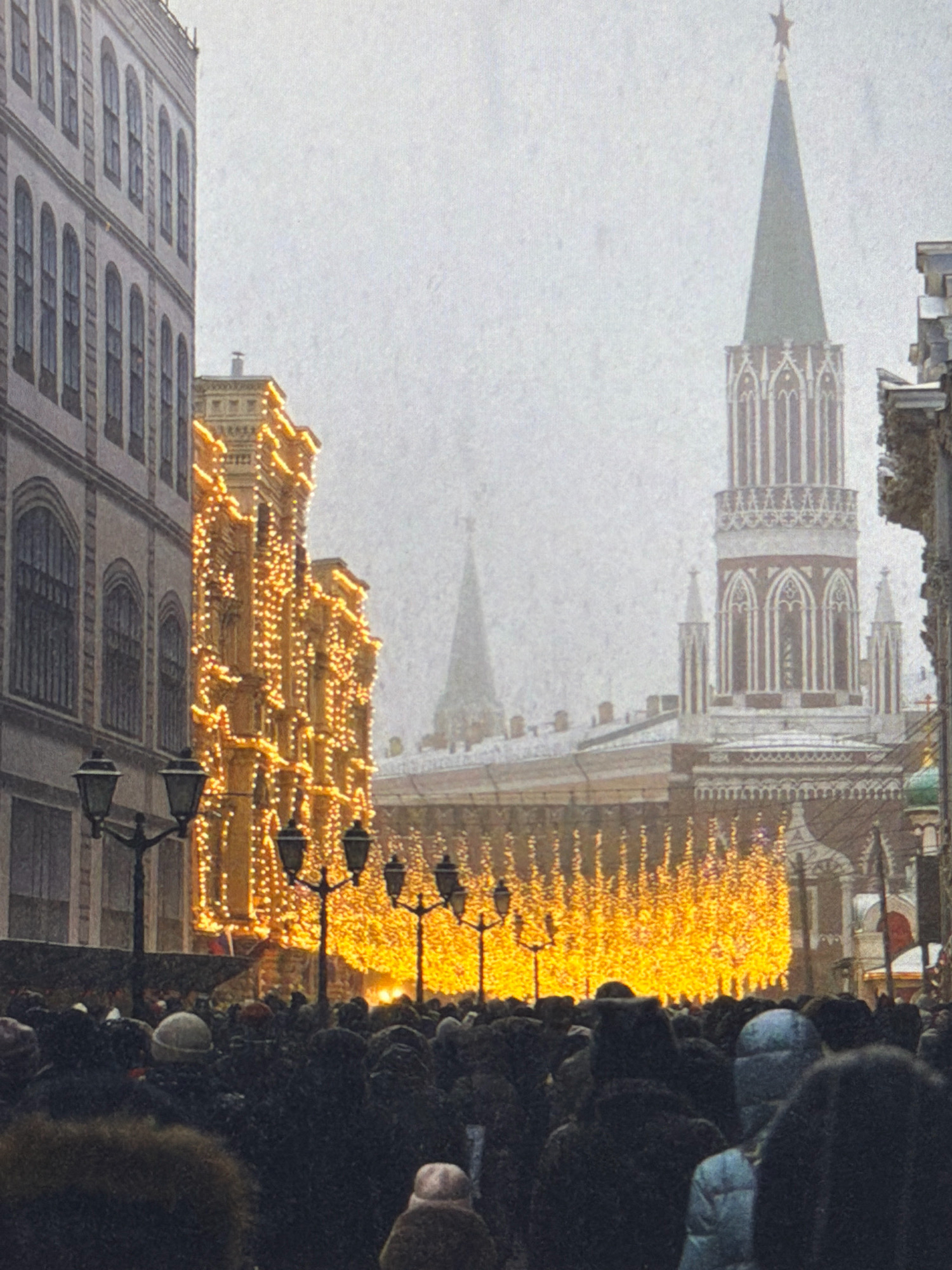 This image depicts a crowded street scene, likely during winter due to the presence of snow or falling precipitation and the heavy winter clothing worn by the people. The architecture suggests a historical European city. On the left, a multi-story building with arched windows and classical architectural details is visible. Adjacent to it, another building is heavily decorated with bright, warm-toned Christmas lights, creating a dazzling display that cascades down its facade.
In the background, a prominent spire, likely part of a cathedral or significant landmark, rises into the hazy sky, topped with a red star. Another, smaller spire can be seen to the left of the main spire. The street is filled with a dense crowd of people, mostly obscured by their winter attire, suggesting a festive or event-filled occasion. Several street lamps with multiple lights are also present, adding to the ambiance. The overall impression is one of a vibrant, illuminated urban environment amidst cold weather.
The image does not depict Kafr Yasif, Israel. The architectural style and the presence of a prominent spire with a red star are characteristic of cities like Moscow, Russia, particularly Red Square during the holiday season.