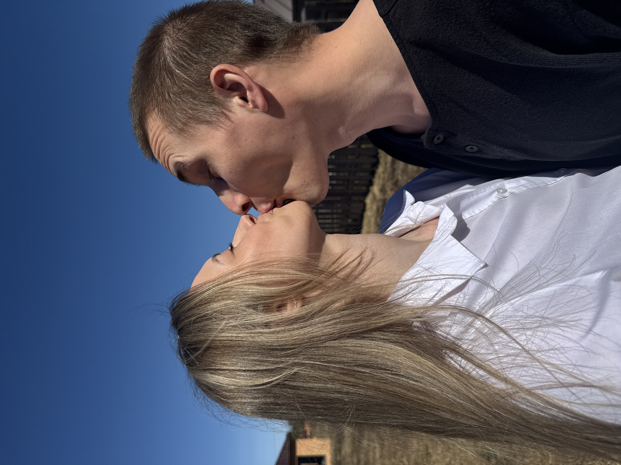 A photo taken outdoors depicts a man and a woman kissing. The woman has long blonde hair and is wearing a white shirt. The man has short brown hair and is wearing a dark shirt. They are positioned in close proximity, with their lips touching. The background includes a clear blue sky and blurred elements suggesting a natural or urban environment. The photo is taken at a low angle, with the subjects angled against the sky. The location may be Chaikovsky, Russia, given the context.