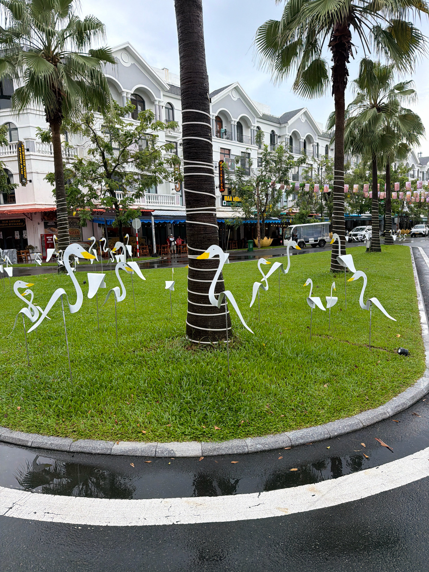 The image depicts a street scene in Phu Quoc, Vietnam, with a focus on a circular grassy median planted with numerous white, abstract sculptures resembling birds. These sculptures, with yellow accents on their beaks, are arranged in a playful manner on slender metal stakes. A large palm tree with white stripes wrapped around its trunk stands prominently in the foreground. In the background, a row of modern, European-style buildings with white facades and grey roofs line the street. The street itself is wet, indicating recent rain, with reflections visible in puddles. A white line painted on the dark asphalt marks the edge of the road. Further down the street, several parked cars and a white van are visible. Decorative banners or flags are strung between palm trees in the middle ground, adding a festive touch.