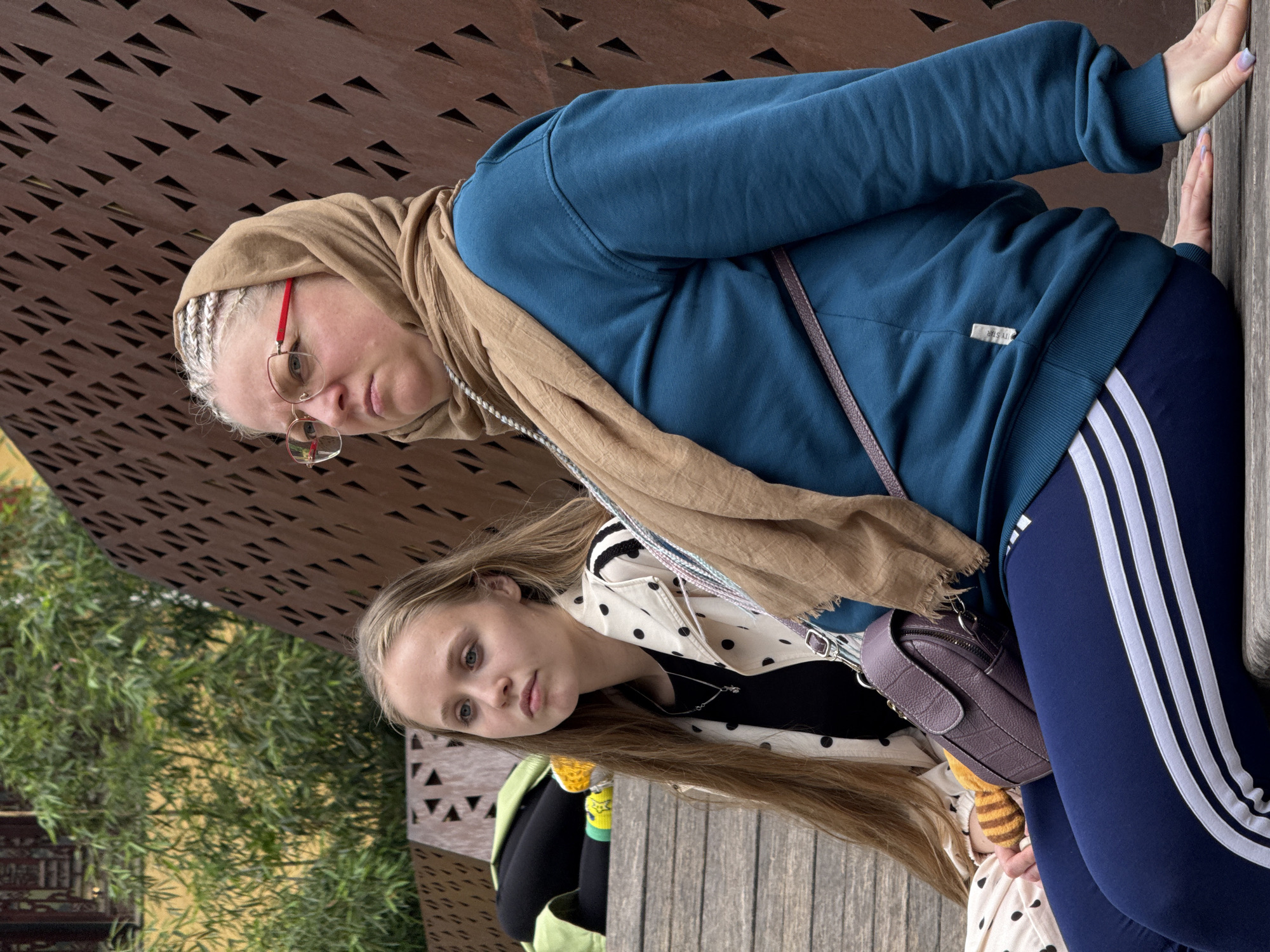 This portrait features two females, likely a mother and daughter, posed outdoors in Yuzhong District, China, during daytime. The image shows the subjects looking directly at the camera with neutral expressions, appearing to be seated or leaning on a wooden structure.
The older woman, positioned on the right side of the frame, wears a light brown headscarf, a dark teal blue hooded sweatshirt, and dark blue athletic leggings with three prominent white stripes on the side. Her blonde and grey hair, styled in braids, is visible beneath the scarf, and she wears distinctive red-framed glasses. A small metallic tag on her sweatshirt reads "MY STR".
The younger girl, on the left, has long, straight light brown hair. She is dressed in a white jacket adorned with black polka dots, worn over a black top, and a delicate silver necklace. A purple textured handbag hangs across her body, and a glimpse of a green garment is visible near her waist.
The background includes a distinctive architectural feature: a brown metallic screen or paneling with numerous small triangular perforations, casting geometric shadows. Behind the girl, lush green foliage, possibly bamboo, adds a natural element to the urban setting. The soft, diffused lighting suggests an overcast day or a shaded area.