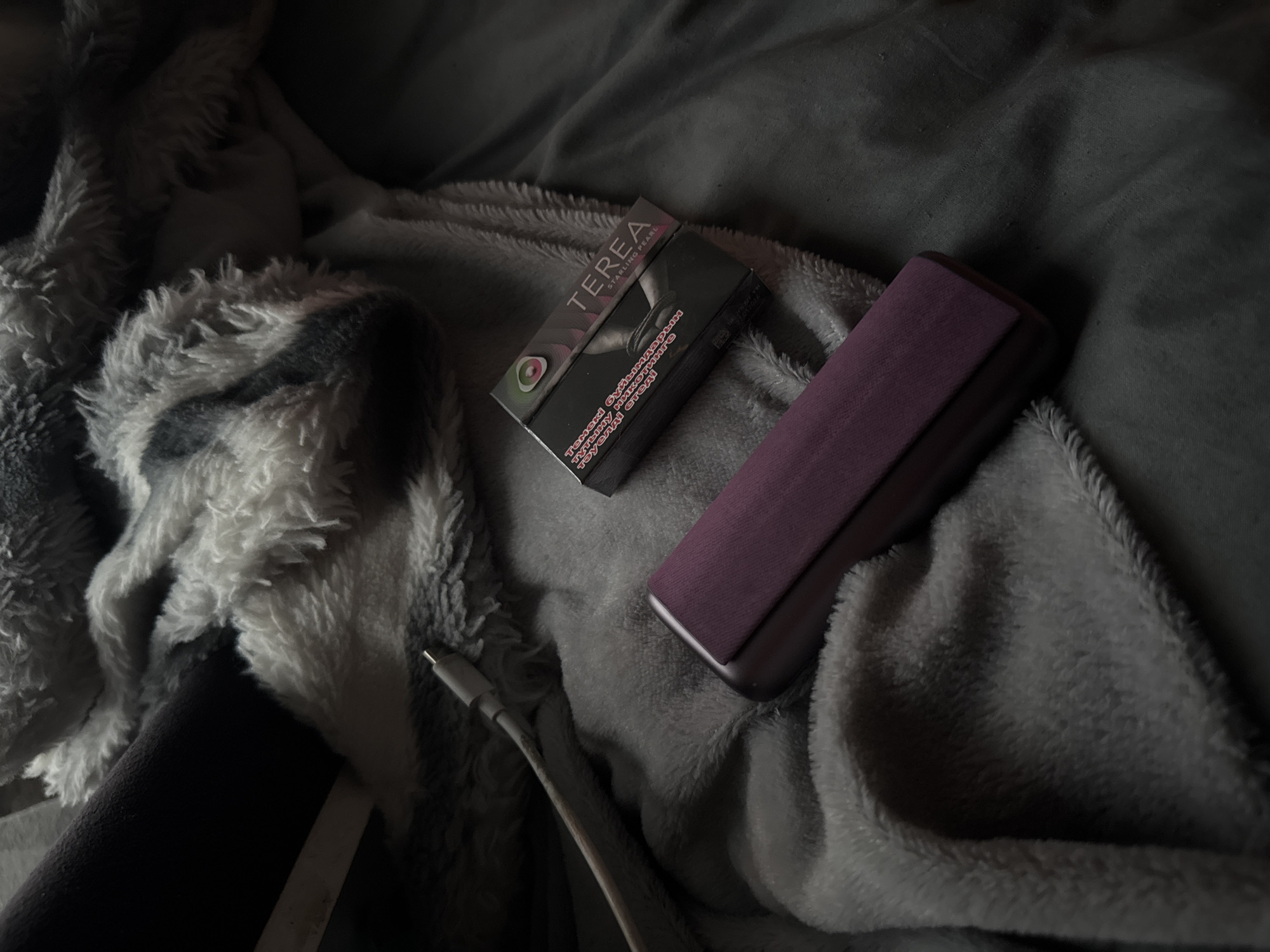 The image shows a close-up, overhead view of a dark-colored, rectangular electronic device with a purple fabric insert, resting on a grey textured surface. To the right of the device is a small, dark cardboard box with colorful branding that reads "TEREA STARLING PEARL". A white charging cable is partially visible in the upper left quadrant of the image, extending diagonally across the scene. The background consists of a plush, grey and white patterned blanket. The lighting is dim, creating shadows and highlighting the textures of the objects. The scene appears to be indoors, on a soft surface, possibly a bed or couch.