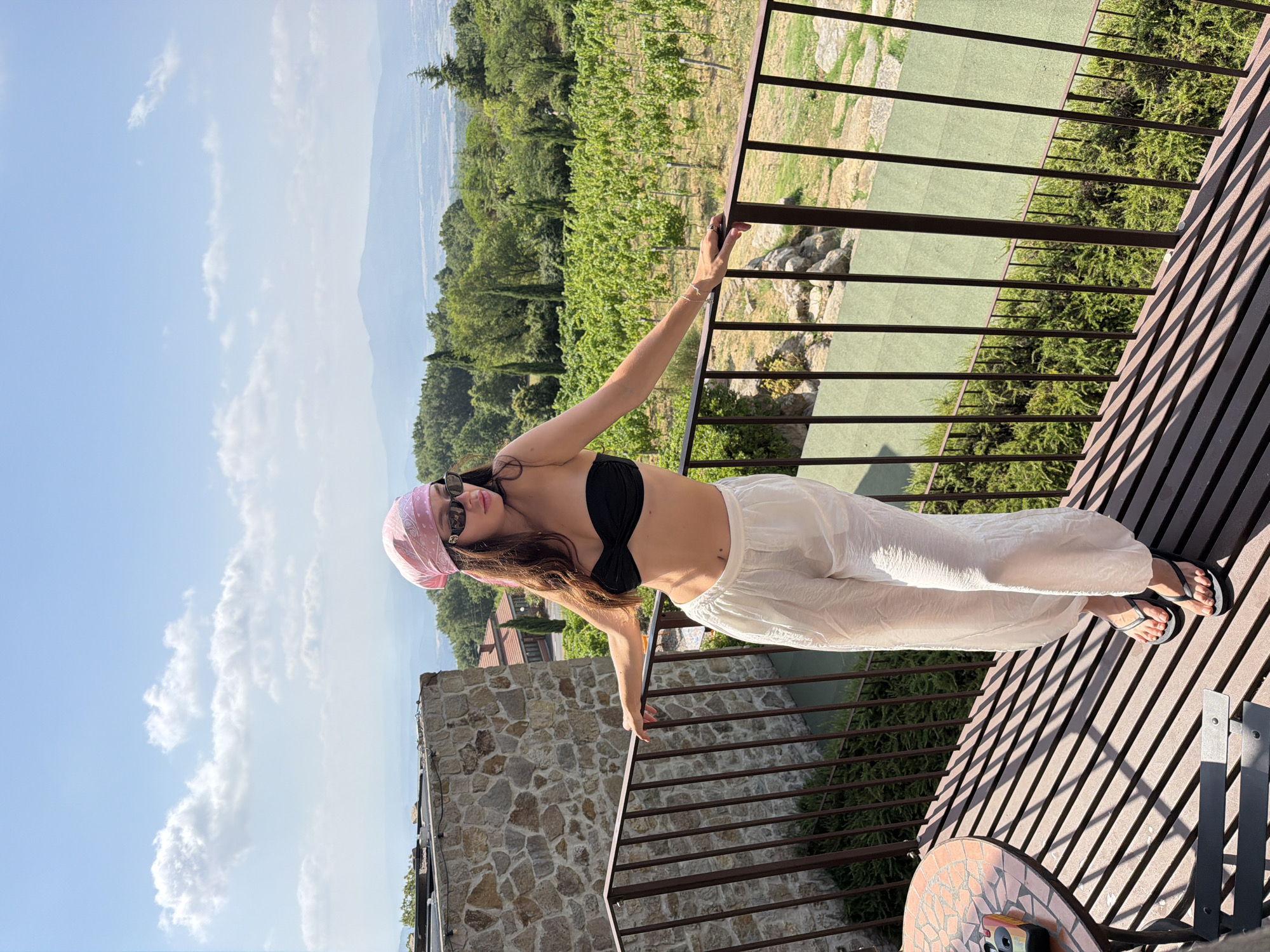 A young woman stands on a balcony overlooking a vineyard. She is wearing a black bikini top, white linen pants, a pink bandana on her head, and sunglasses. The balcony has a metal railing and a table with a small camera. In the background, there are rolling hills covered in vineyards and trees. The sky is blue with scattered clouds. The location is a vineyard in Sassofortino, Italy.