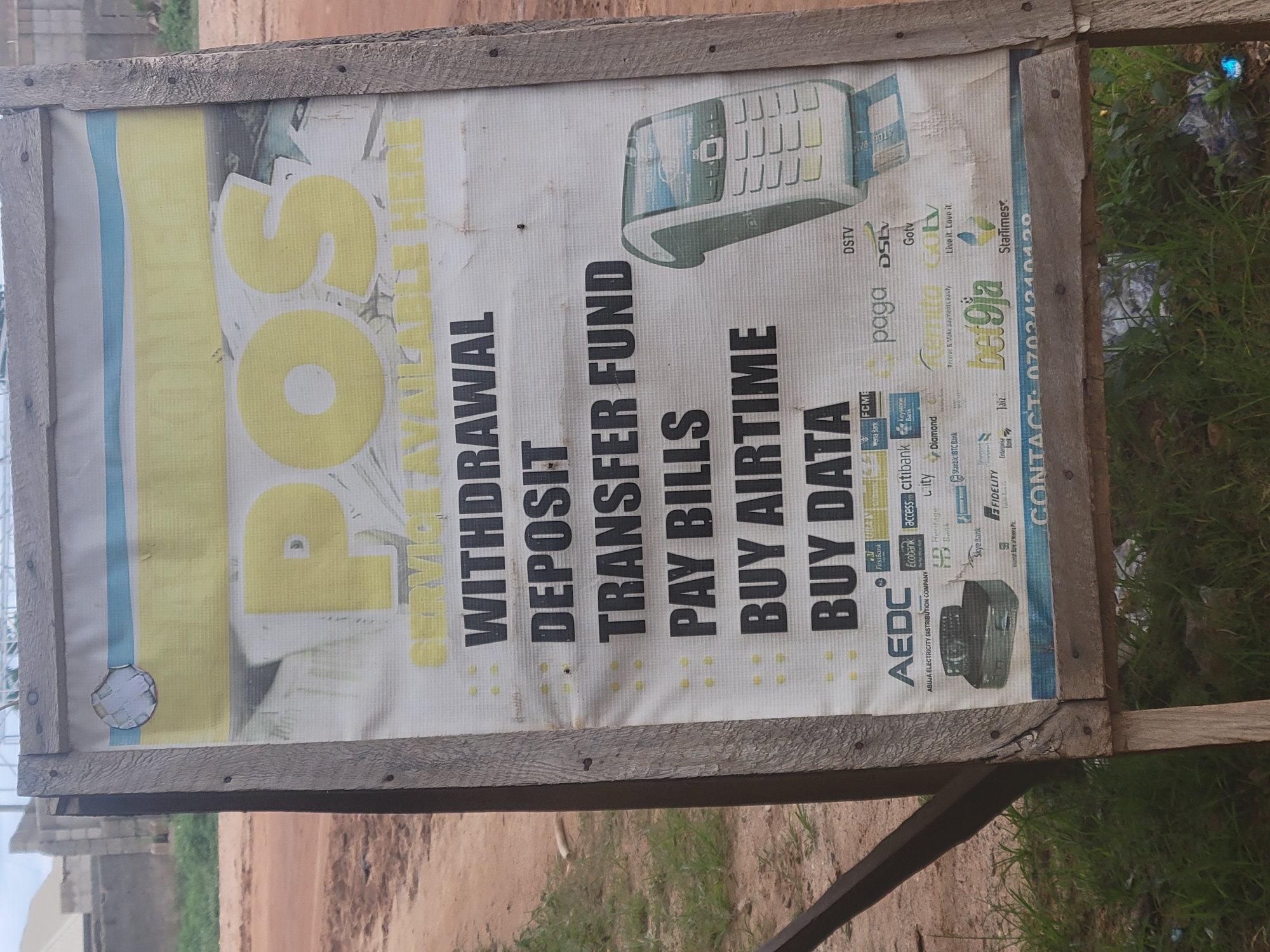 An outdoor, weathered wooden signboard stands angled to the viewer's right, displaying a white vinyl banner in Keffi, Nasarawa State, Nigeria. The signboard frame is constructed from rough, light-brown wooden planks joined by visible nails. The banner advertises "POS SERVICE AVAILABLE HERE," with "POS" prominently displayed in large yellow letters across the upper middle section. Below this, a list of services is presented in black, bold text with small yellow square bullet points: "WITHDRAWAL," "DEPOSIT," "TRANSFER FUND," "PAY BILLS," "BUY AIRTIME," and "BUY DATA."
On the right side of the banner, a graphical depiction of a grey-green mobile Point of Sale (POS) terminal with a keypad and screen is visible, featuring a blue credit card partially inserted into its slot. A second, older-model, dark grey POS terminal graphic is positioned in the bottom left. The lower section of the banner is covered with a collage of logos from various Nigerian financial institutions, payment platforms, utility providers, and entertainment services, including AEDC (Abuja Electricity Distribution Company), FirstBank, Ecobank, Access Bank, Fidelity Bank, Union Bank of Nigeria Plc., Paga, Remita, Bet9ja, DSTV, GOtv, and Startimes. Partially visible contact information, "CONTACT: 07024210420," is at the very bottom. The banner shows signs of wear, including creasing and a circular tear in the upper left corner.
The immediate foreground consists of dry, reddish-brown soil interspersed with sparse green grass. A small piece of crumpled blue plastic litter is visible among the grass in the bottom right corner. In the mid-ground, a dusty, reddish-brown dirt road extends horizontally across the left side of the frame, disappearing into the background where simple, unpainted concrete block buildings and scattered green foliage are visible under a bright, diffused sky.