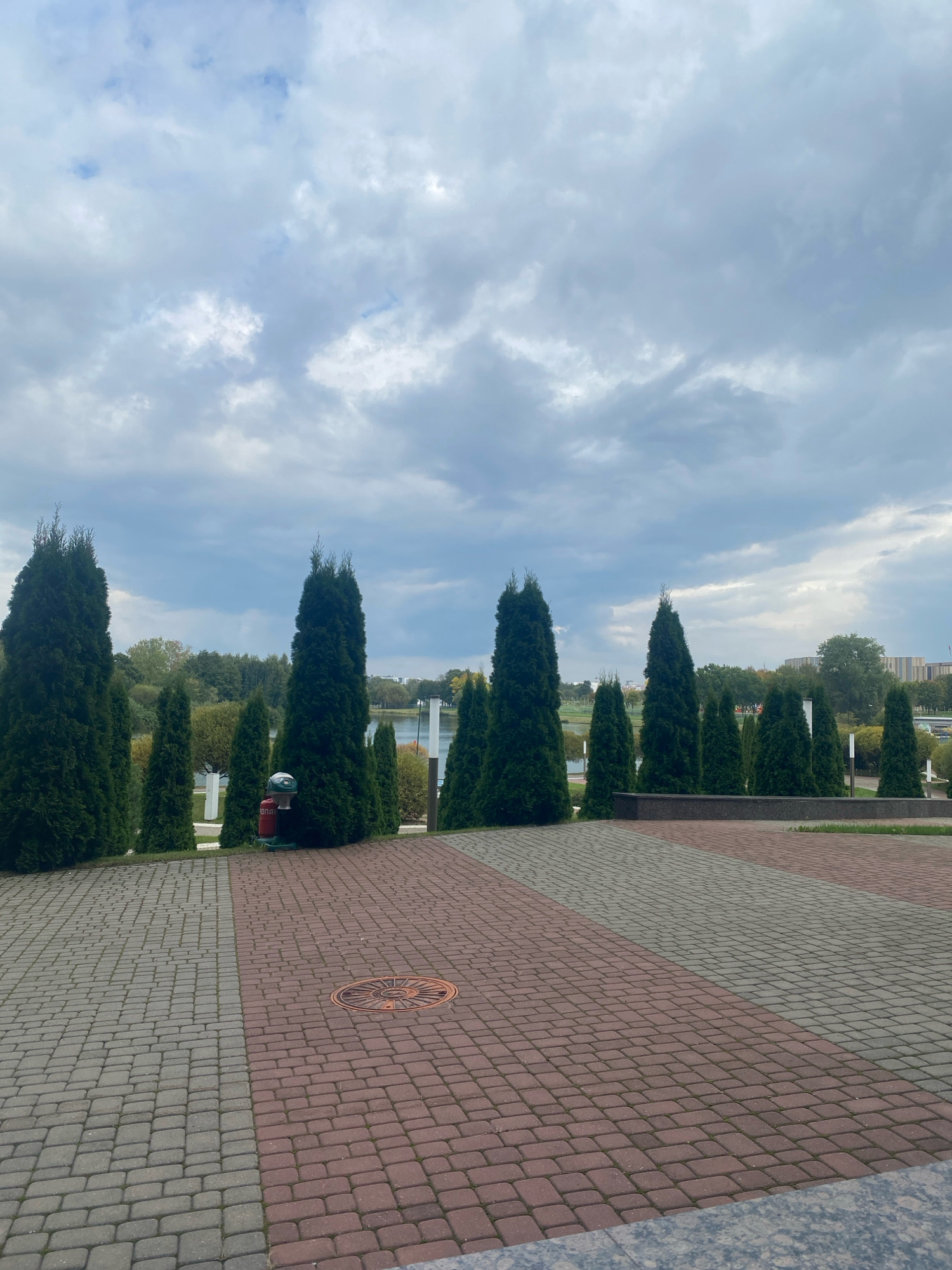 An overcast day is depicted with a view of a park area. In the foreground, a paved pathway is laid with two contrasting colors of rectangular stones: grey on the left and reddish-brown on the right. A circular metal manhole cover with an ornate design is visible in the reddish-brown section of the path.
A line of tall, slender evergreen trees forms a backdrop to the pathway, with some smaller trees and bushes interspersed. To the left of the trees, a red fire hydrant with a silver top stands out. Beyond the trees, a body of water is visible, likely a lake or river, with a distant treeline on the other side. In the far background, buildings and more trees are scattered across the horizon.
The sky is filled with a thick layer of grey and white clouds, suggesting a cloudy or potentially rainy day. The overall scene suggests a public park or recreational area within the city of Minsk, Belarus, designed for walking and relaxation.
