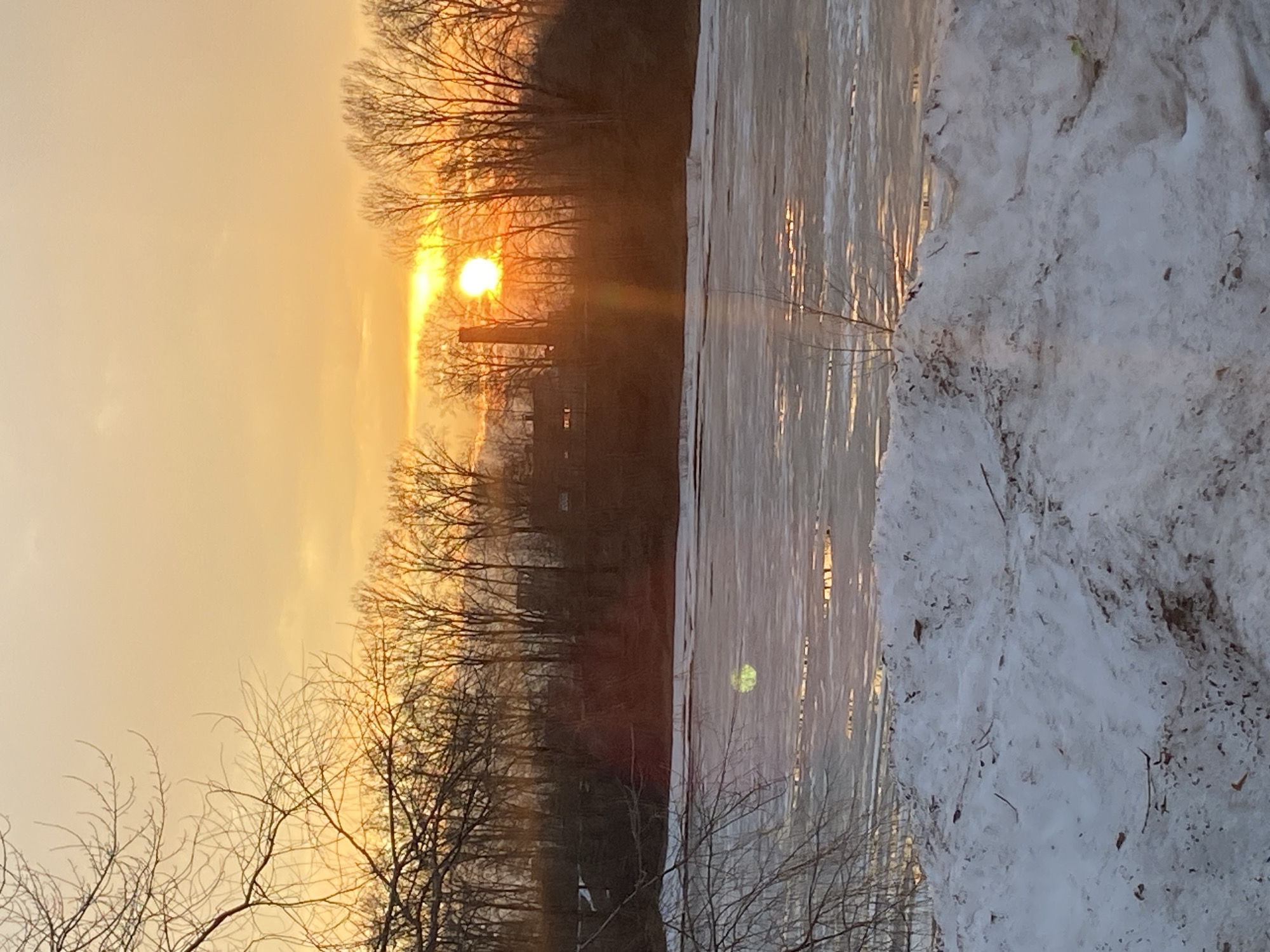 The image shows a winter scene, potentially at sunset or sunrise, with a body of water largely covered in ice and snow. The sky is a yellowish-orange hue, with the sun visible through bare trees on the far shore. There appears to be some industrial structure visible. The right foreground features a bank of snow with sparse vegetation. It cannot be ascertained the location of Chaikovsky in Russia.