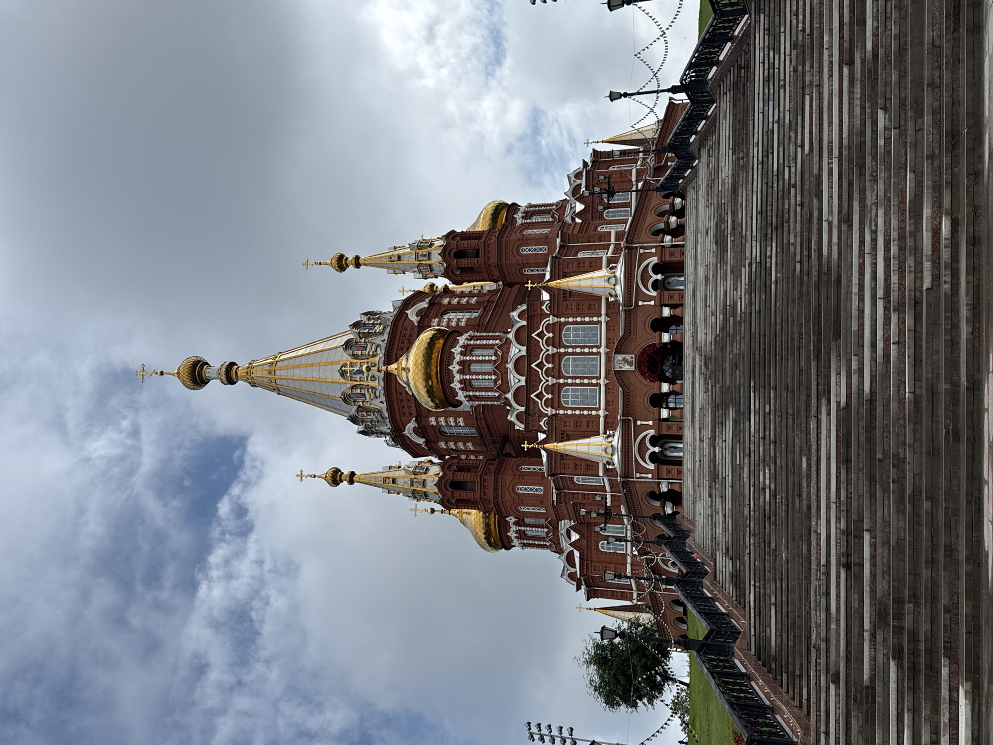 The image shows the Alexander Nevsky Cathedral in Izhevsk, Russia, viewed from a wide, long staircase. The cathedral is a large, red brick building with multiple golden domes and intricate white architectural details. It is topped with crosses. The sky above is cloudy. The staircase is made of stone or concrete, with dark metal railings on either side, leading up to the entrance of the cathedral. There are streetlights and decorative string lights along the railings. A few trees are visible on the grassy slope to the left of the stairs. There are no people visible in the foreground or on the stairs.