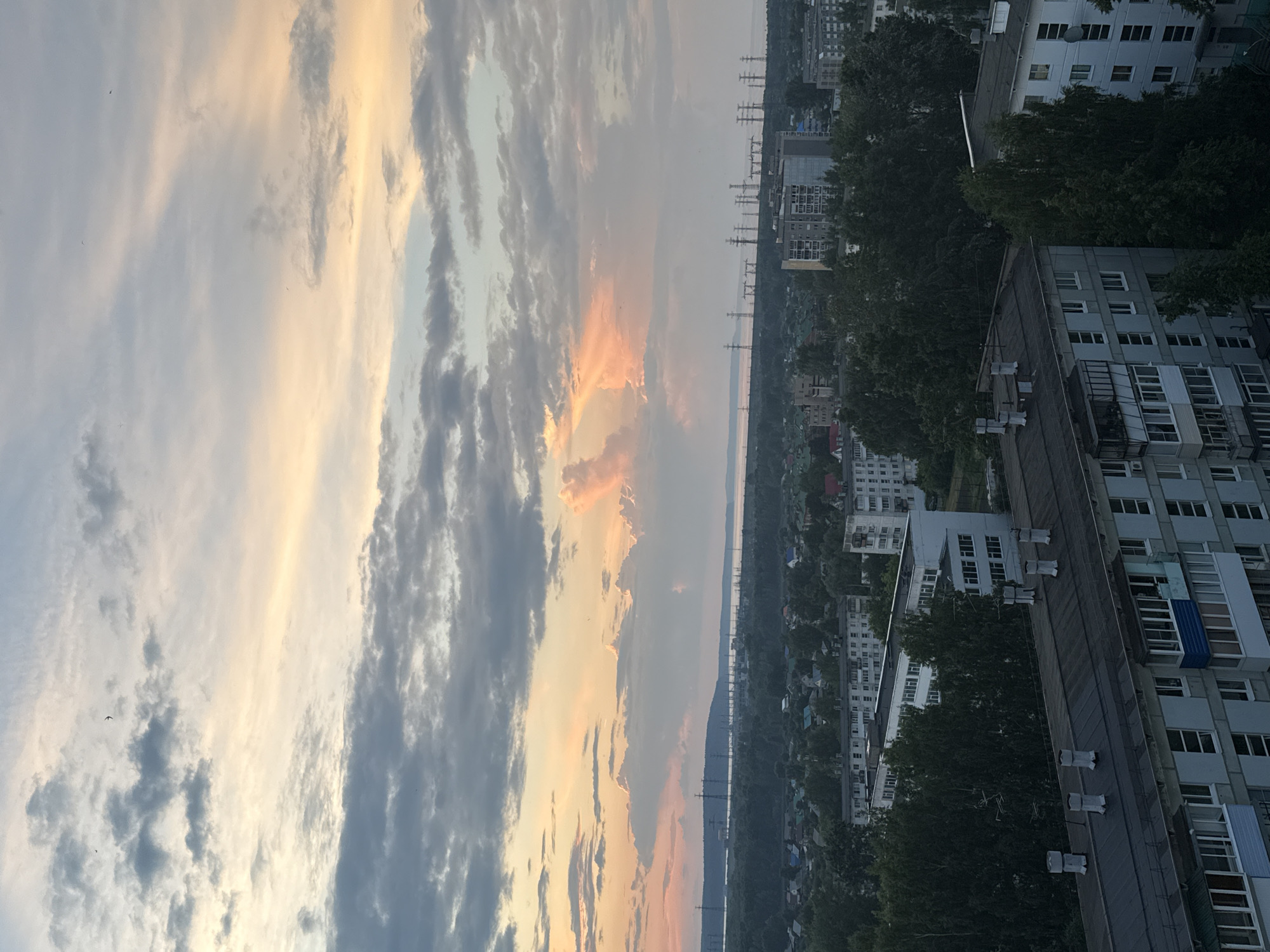 An aerial perspective captures a cityscape under a dramatic sky during what appears to be sunset or sunrise. The sky is filled with dynamic clouds, with patches of soft orange and yellow hues indicating the presence of the sun behind or between them, contrasting with darker gray clouds. In the distance, a body of water stretches across the horizon, with a line of tall electrical pylons visible along its banks and extending inland.
The middle ground reveals a densely wooded area interspersed with buildings, suggesting a transition from rural to urban development. Numerous residential buildings, likely apartment blocks, are prominent in the foreground. These structures are multi-story, with visible windows and balconies. Some buildings have flat, dark roofs with visible exhaust vents or chimneys. The architecture suggests a post-Soviet era construction style. Trees, mostly deciduous, are abundant throughout the scene, particularly in the mid-ground and surrounding the buildings. The overall impression is of a large settlement situated near a natural landscape, under a vibrant and cloudy sky.