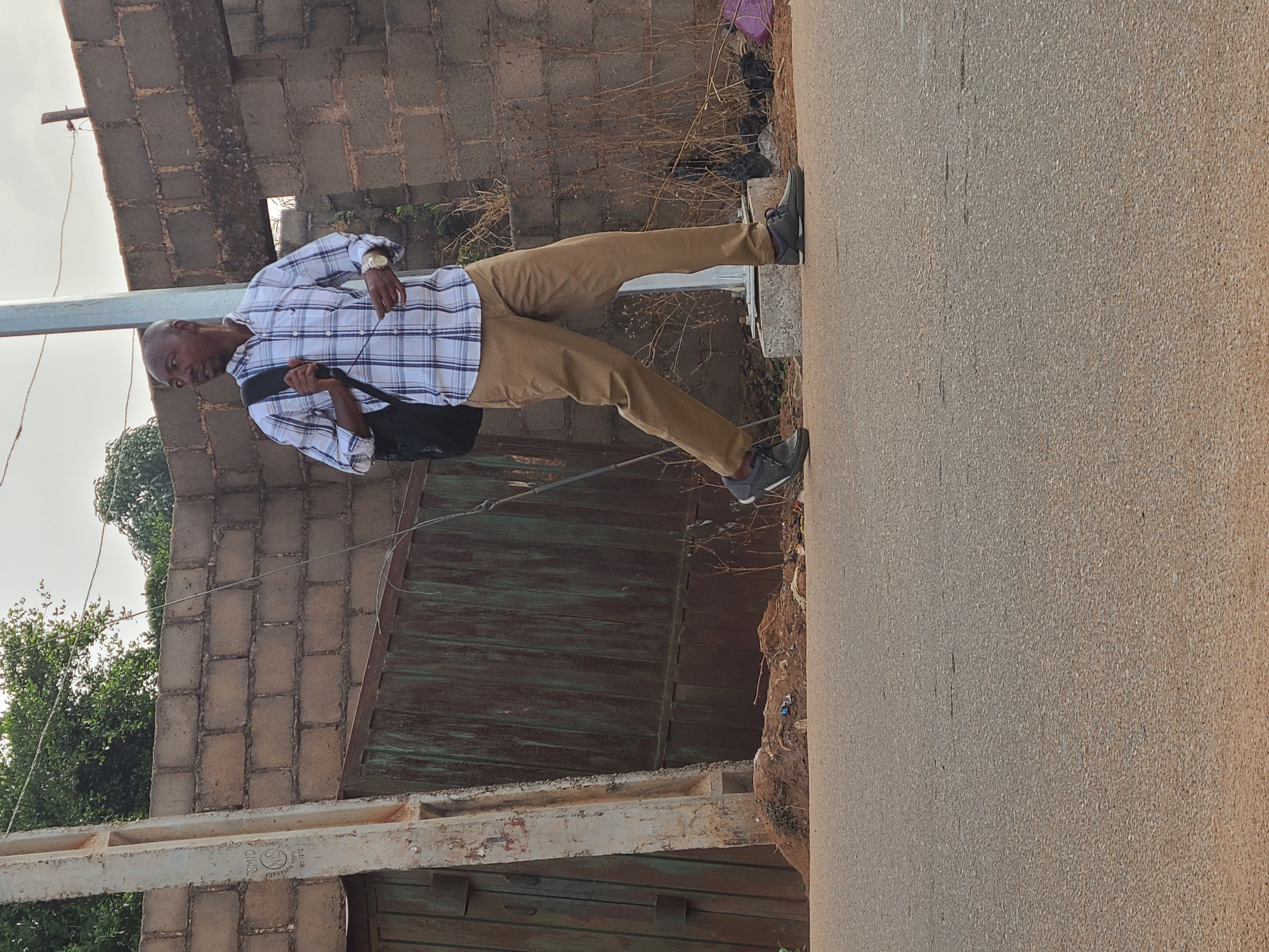 A man stands on the side of a paved road in Jos, Nigeria. He is wearing a plaid shirt, khaki pants, and sneakers. He has a black bag slung over his shoulder. Behind him are a brick wall, a gate, vegetation, and utility poles with wires. The road appears to be asphalt and has an orange-brown hue from the sunlight.