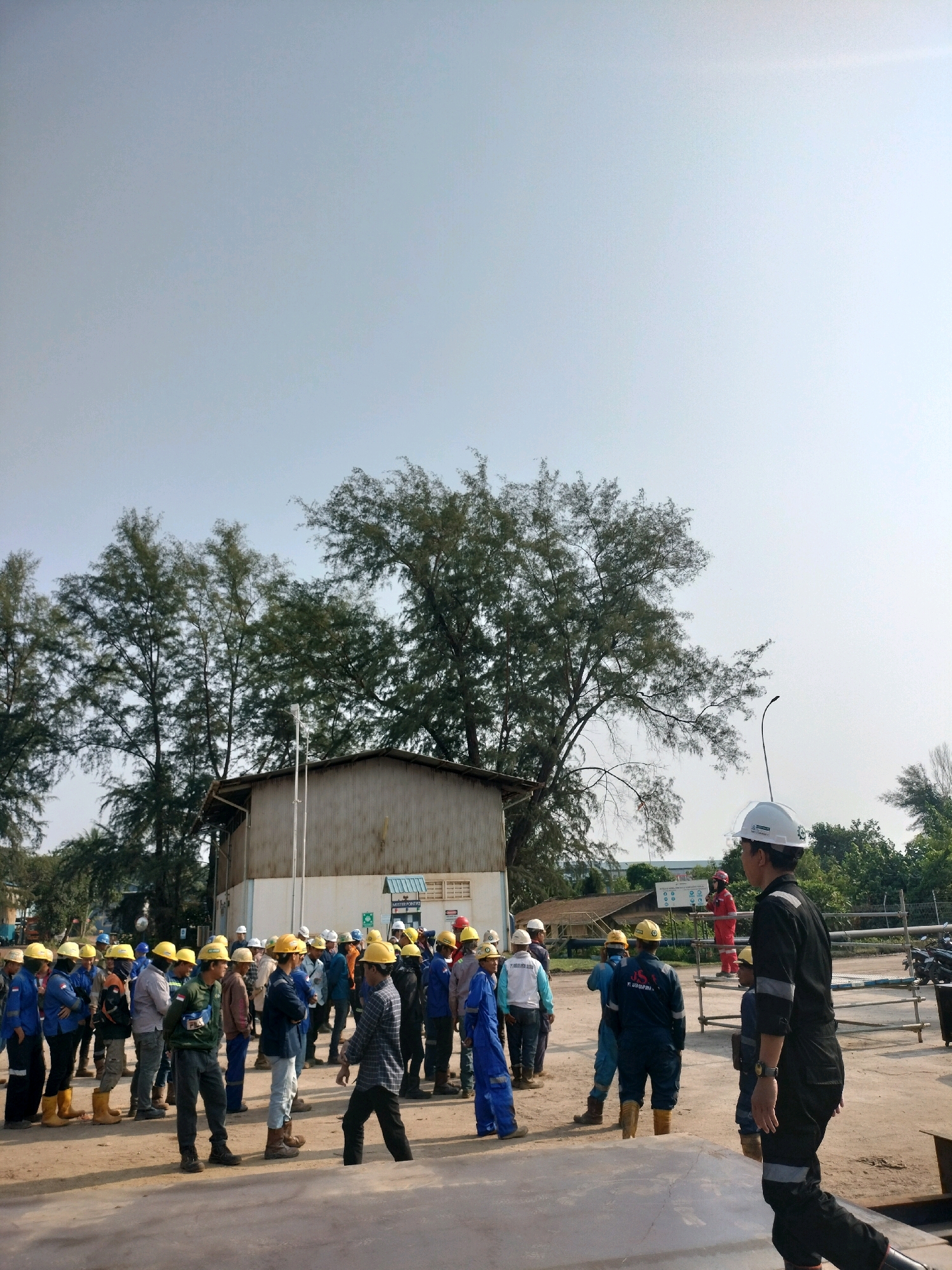 The image depicts an outdoor scene at what appears to be an industrial site or construction area in Batam, Indonesia. A group of approximately 20-30 individuals, most of whom are wearing hard hats and work uniforms, are gathered in an open, dusty area. The individuals are of varying ages and ethnicities and appear to be engaged in a briefing or instruction session. Some are standing and listening, while others are in motion, possibly moving to or from the group.
In the background, there is a long, low-slung building with a corrugated metal exterior and roof, suggesting it might be a workshop or storage facility. Several mature trees with dense foliage are visible behind the building, and the sky is clear and bright, indicating daytime. There are also some scaffolding structures and industrial equipment present, further reinforcing the industrial setting.
One individual in the foreground, on the right side of the image, is wearing a black jumpsuit with reflective stripes and a white hard hat. This person is facing away from the camera and walking, possibly towards or away from the group. Another person, in a red jumpsuit and white hard hat, is standing on an elevated platform in the mid-ground, near a sign that reads "MASTER POINTER." The presence of hard hats and uniforms strongly suggests a work environment where safety protocols are in place. The overall atmosphere appears to be one of an active work site.