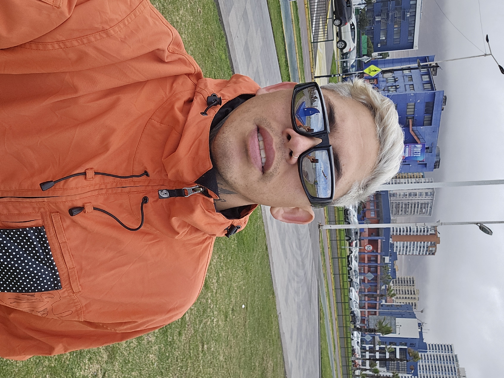 A man with dyed blonde hair and sunglasses is taking a selfie outdoors. He is wearing an orange hooded jacket. In the background, there are several modern buildings, some of which are blue and white. There are cars parked along a street and a grassy area in the foreground. The scene appears to be in a coastal city, likely along the waterfront promenade in Iquique, Chile, suggested by the architecture and the presence of a body of water visible in the reflection of the man's sunglasses. The sky is overcast, and the lighting is diffused.