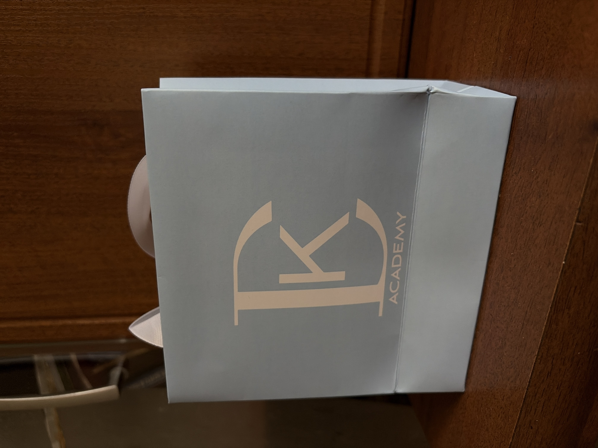 A light blue paper gift bag with a peach-colored logo of a stylized "DK" and the word "ACADEMY" below it sits on a dark brown wooden surface, possibly a table or shelf. The bag has white ribbon handles, with a portion of one handle visible at the top left. The background is out of focus, but appears to be a wooden cabinet or wall. The scene is indoors.