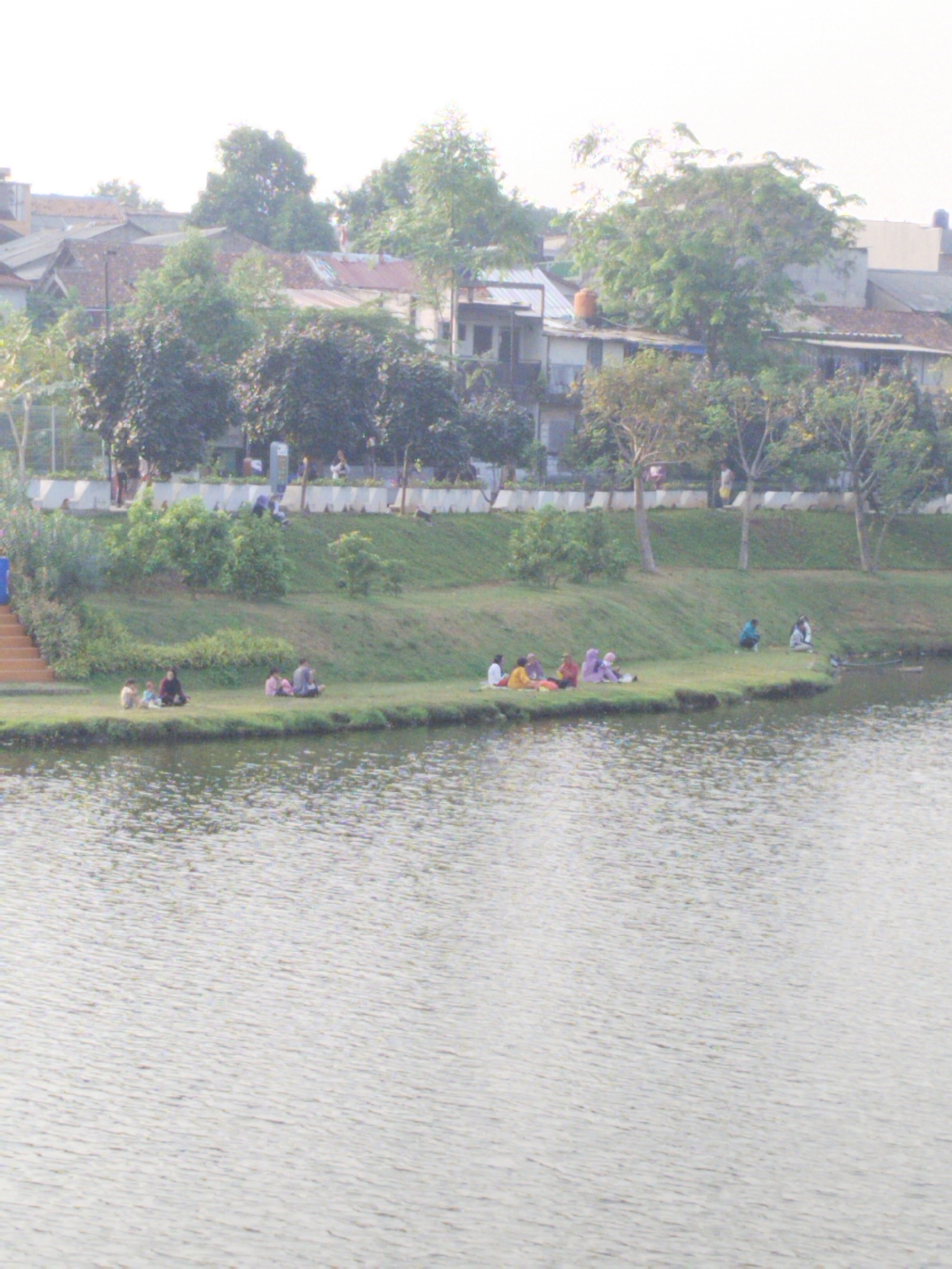 The image depicts a serene lakeside scene in Depok, Indonesia. A group of people are enjoying a picnic on the grassy bank of a calm body of water. Several individuals are seated together, some appearing to be families with children, engaged in conversation and relaxation. The background reveals a lush green environment with trees and vegetation, bordering a residential area with buildings and houses. The atmosphere suggests a peaceful day outdoors, with people embracing nature and community.