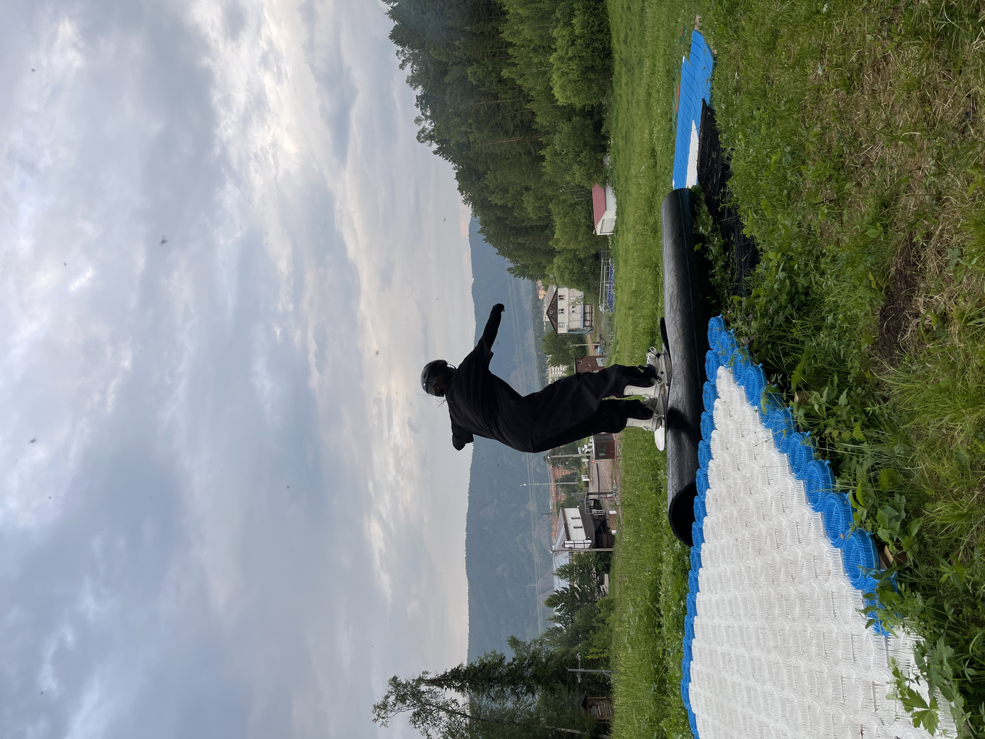 This image shows a person in a black outfit and helmet standing on a large black mat with a white and blue patterned surface. The person is in a dynamic pose, with one arm extended upwards and the other bent at the elbow. The mat appears to be part of a recreational setup, possibly for activities like gymnastics or sports.
The scene is set outdoors, with lush green grass and trees surrounding the mat. In the background, there are buildings and a vast mountain range under a cloudy sky. The lighting suggests it's either early morning or late afternoon. The overall atmosphere is somewhat serene, with the natural landscape providing a beautiful backdrop to the activity.
There is no visible text in the image. The presence of buildings and the mountainous terrain hint at a location near a settlement in a hilly or mountainous region. The weather appears overcast, with dramatic clouds filling the sky. The person's posture and the surrounding environment suggest an active pursuit in a natural setting.