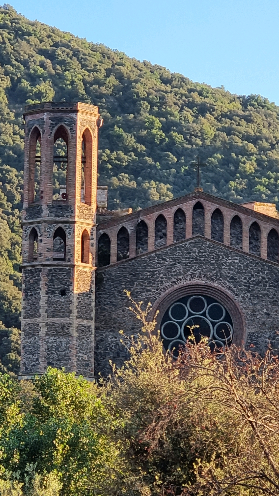 The image shows a close-up, eye-level view of the stone and brick church of Sant Joan Les Fonts, Spain, set against a backdrop of a sunlit, tree-covered hillside. The church features a prominent bell tower on the left, constructed with alternating bands of stone and red brick, with arched openings at the top. Adjacent to the tower is the main facade of the church, also built of stone with arched windows along the roofline. A large circular rose window with multiple smaller circular panes is visible on the facade. The scene is bathed in the warm light of what appears to be late afternoon sun, casting shadows and highlighting the textures of the architecture and the foliage. Green trees and bushes are in the foreground, obscuring the lower part of the church and creating a natural frame for the building. The sky above is clear and blue. No people are visible in the image.