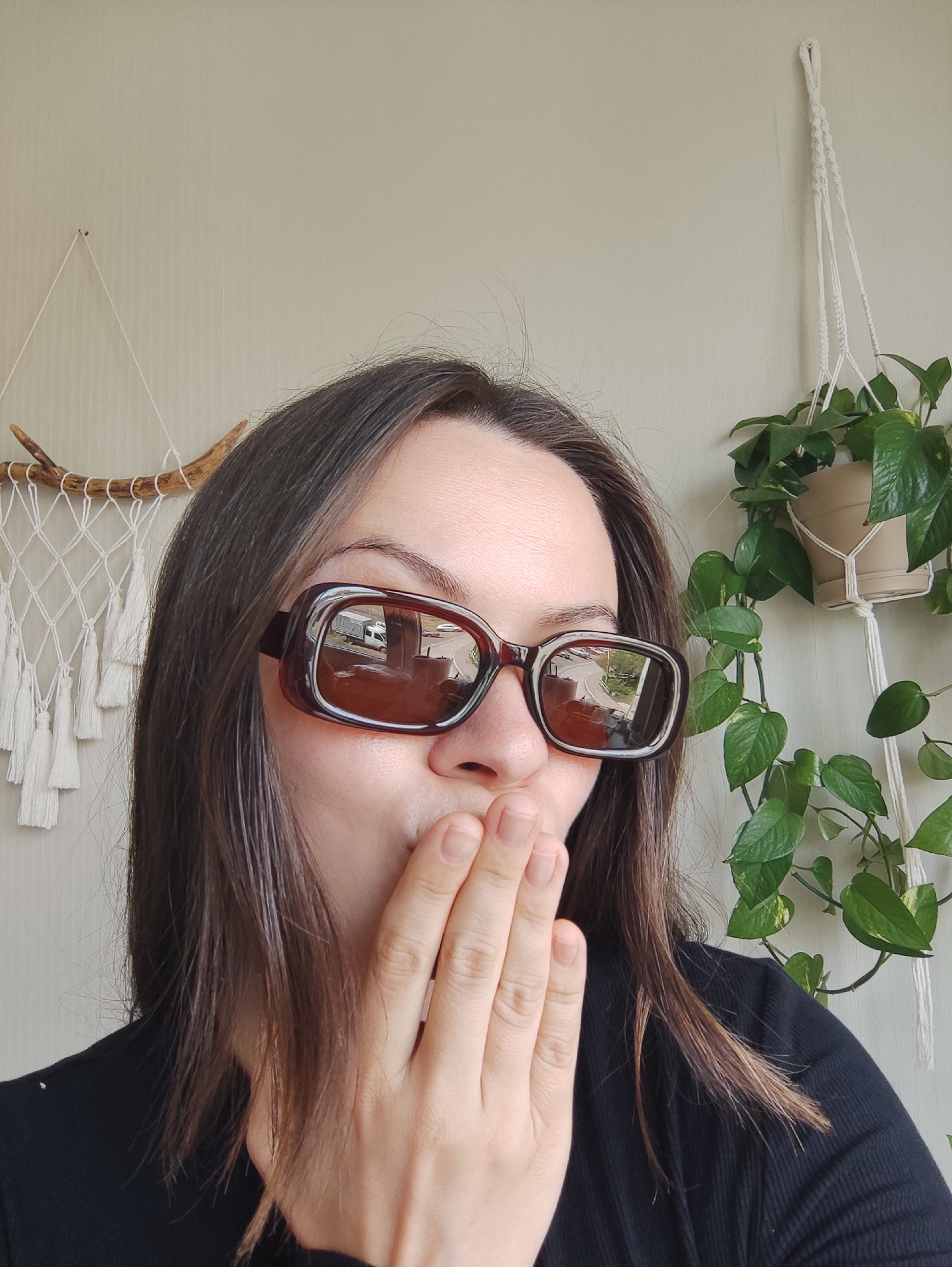 A woman is photographed indoors, wearing large, brown-framed sunglasses. Her hand is raised to her mouth. Her dark hair is straight and shoulder-length. She is wearing a black top. In the reflection of her sunglasses, one can see cars, a truck, and buildings indicating a streetscape. Behind her, a wall is visible, adorned with a macrame wall hanging on the left and a hanging potted plant on the right. Based on the reflection in the sunglasses, the location is identifiable as Chaikovsky, Russia.