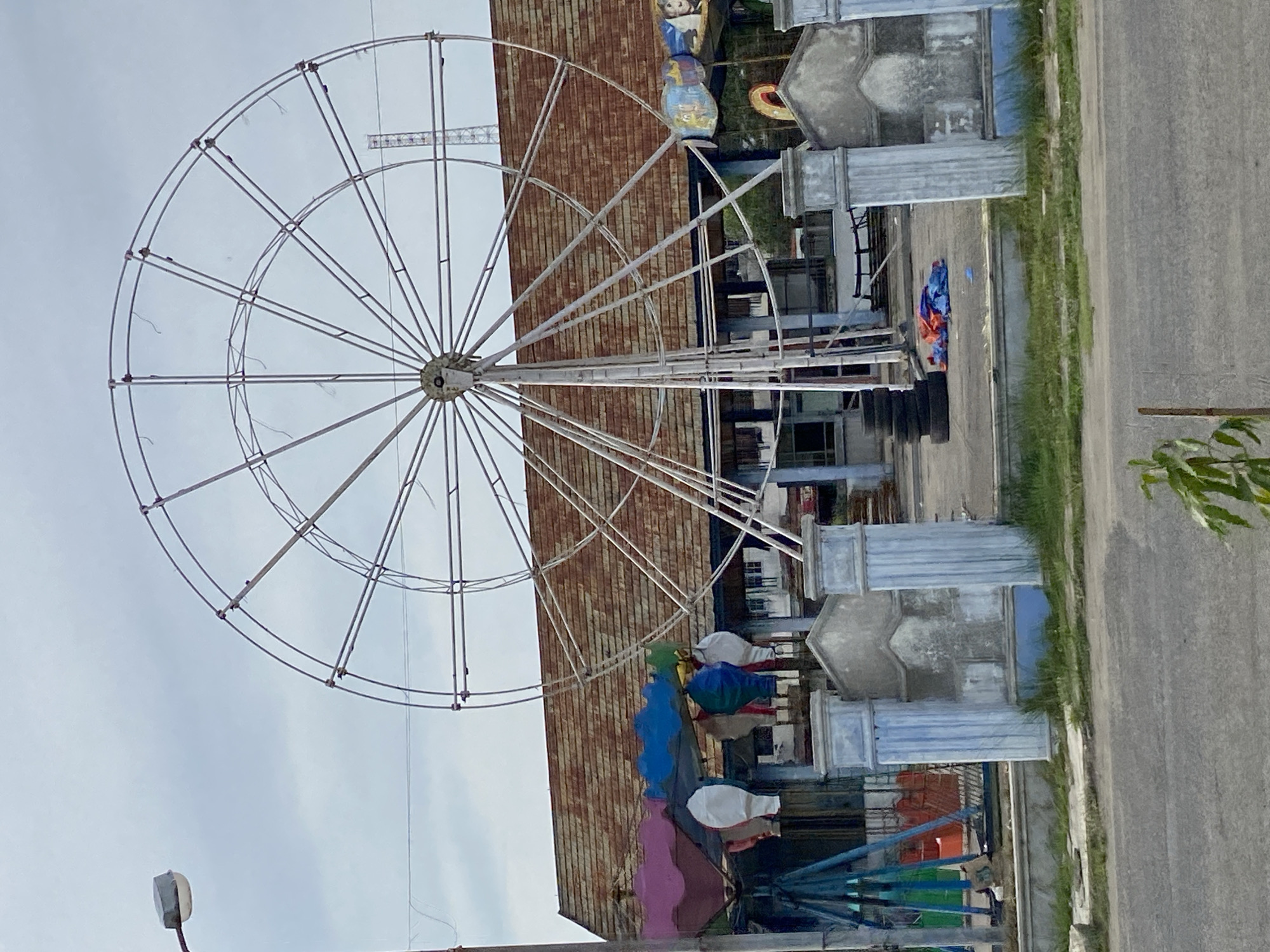 The image depicts a vacant amusement park area in Lhokseumawe, Indonesia. A large, incomplete Ferris wheel dominates the center of the frame, its metal structure stark against an overcast sky. To the left of the Ferris wheel, a carousel with colorful, balloon-like structures stands idle. In front of the carousel, a blue and green swing set is visible. A building with a rusted, corrugated metal roof serves as a backdrop for the attractions.
The ground within the park is paved, with a pile of tires and some discarded tarps situated near the base of the Ferris wheel. A low concrete wall with blue-painted pillars flanks the entrance to the park. The area appears to be in a state of disrepair and abandonment. A street runs across the foreground, with a lone sapling growing on the grassy verge between the road and the park. A streetlamp is positioned to the left of the Ferris wheel, its light fixture covered by a gray cap. In the distance, a tall, lattice-like structure, possibly a communication tower, can be seen. There are no people or animals visible in the scene.