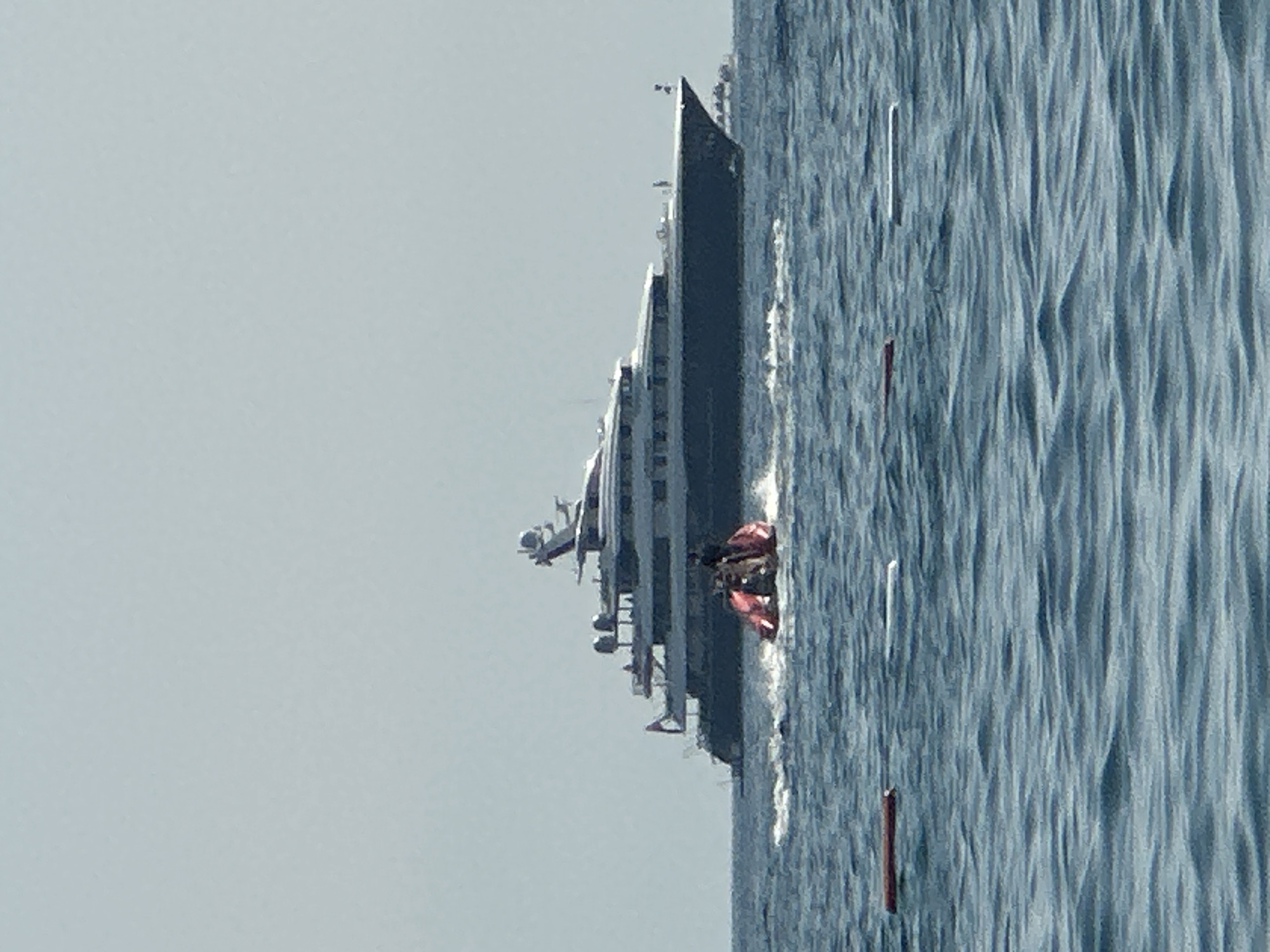 In the waters off Boreti, Montenegro, a large, multi-decked yacht with a dark hull and white superstructure sails on a calm, blue sea under a hazy sky. In the foreground, a red and black personal watercraft with at least two individuals aboard is speeding across the water, creating a wake. Several buoys, some red and some white, are visible floating on the surface of the water. In the distance, the outline of another, smaller vessel is discernible.