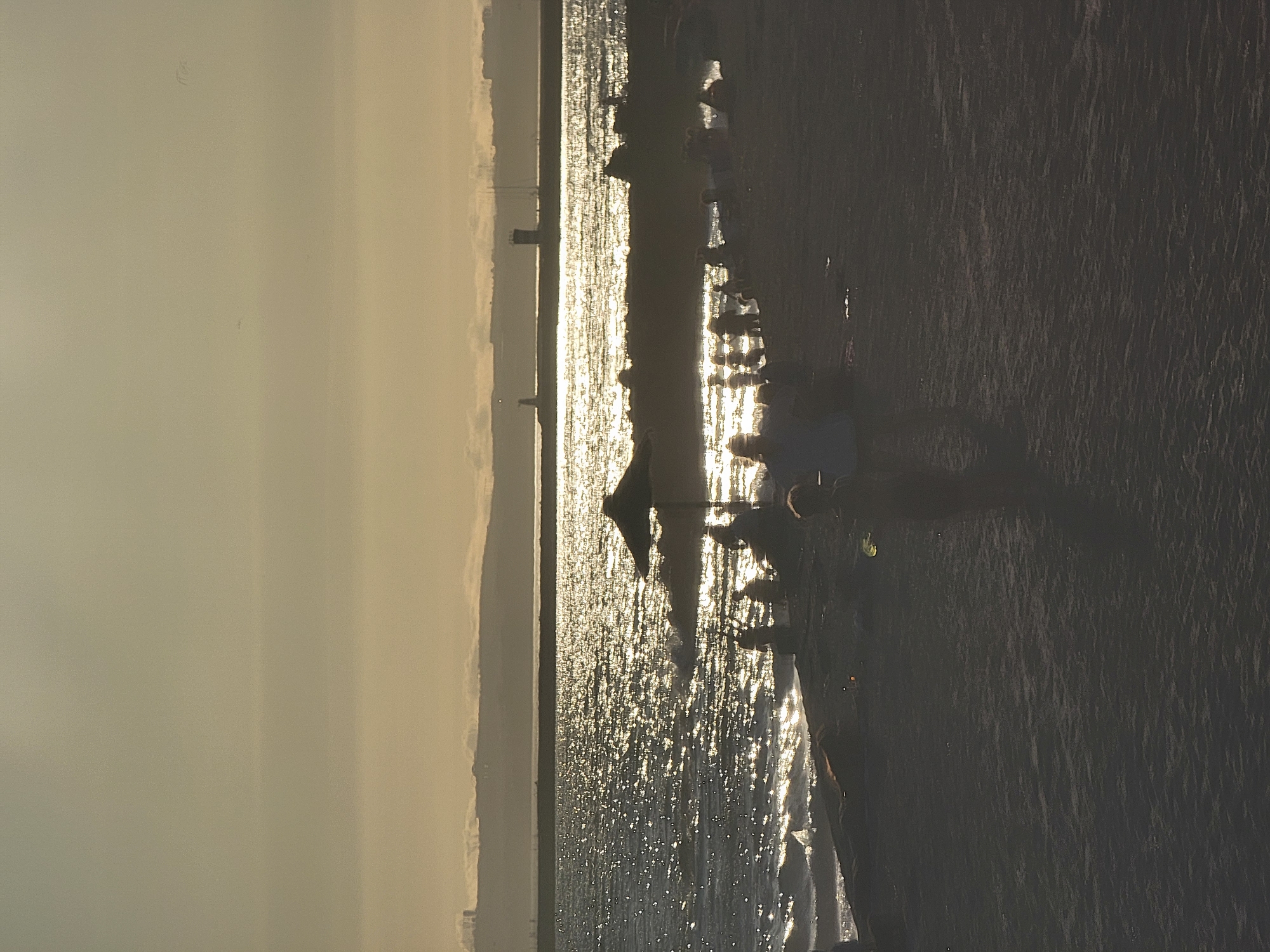 This is a photograph taken from the Quarteira beach in Quarteira, Portugal, during sunset. The sun is low on the horizon, casting a golden shimmer across the sea, creating a sparkling effect on the water's surface. In the foreground, the sandy beach is visible with several silhouetted figures. Some individuals are gathered, while others appear to be walking or standing. One prominent dark umbrella stands on the beach, casting a shadow. In the middle ground, the sea meets the shore with small waves. In the background, a long, low landmass is visible, possibly the edge of the coast or an island, with faint structures that could be buildings or lighthouses on the horizon. The sky is a gradient of light, with the lower portion near the horizon brighter due to the sunset. The overall scene depicts a serene beach environment at dusk.
